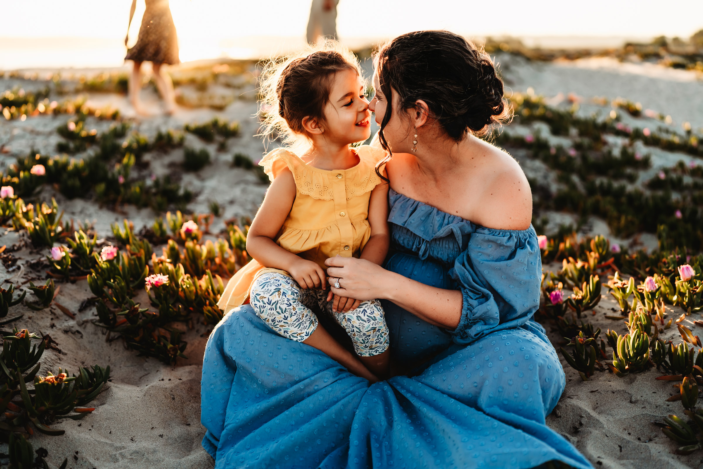 Pregnant woman sitting on a sand dune holding her daughter on her lap during a lifestyle maternity photo session on Coronado Beach.