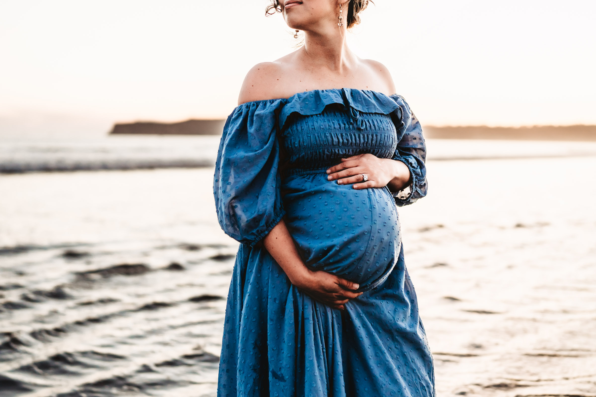 Pregnant woman holding her belly and looking over her shoulder during a lifestyle maternity photo session on Coronado Beach.