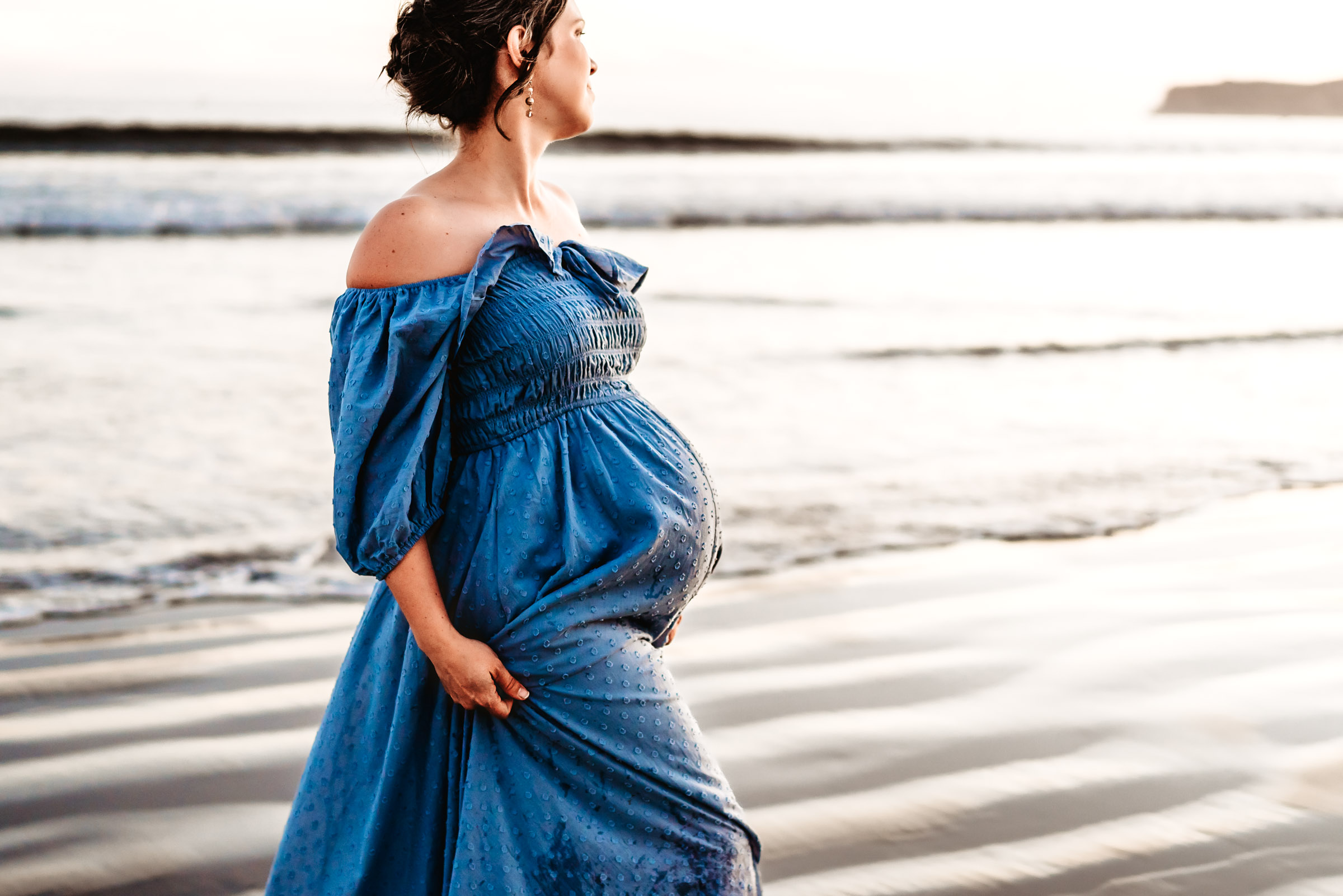 Pregnant woman holding her dress and looking at the beach sunset during a lifestyle maternity photo session on Coronado Beach.