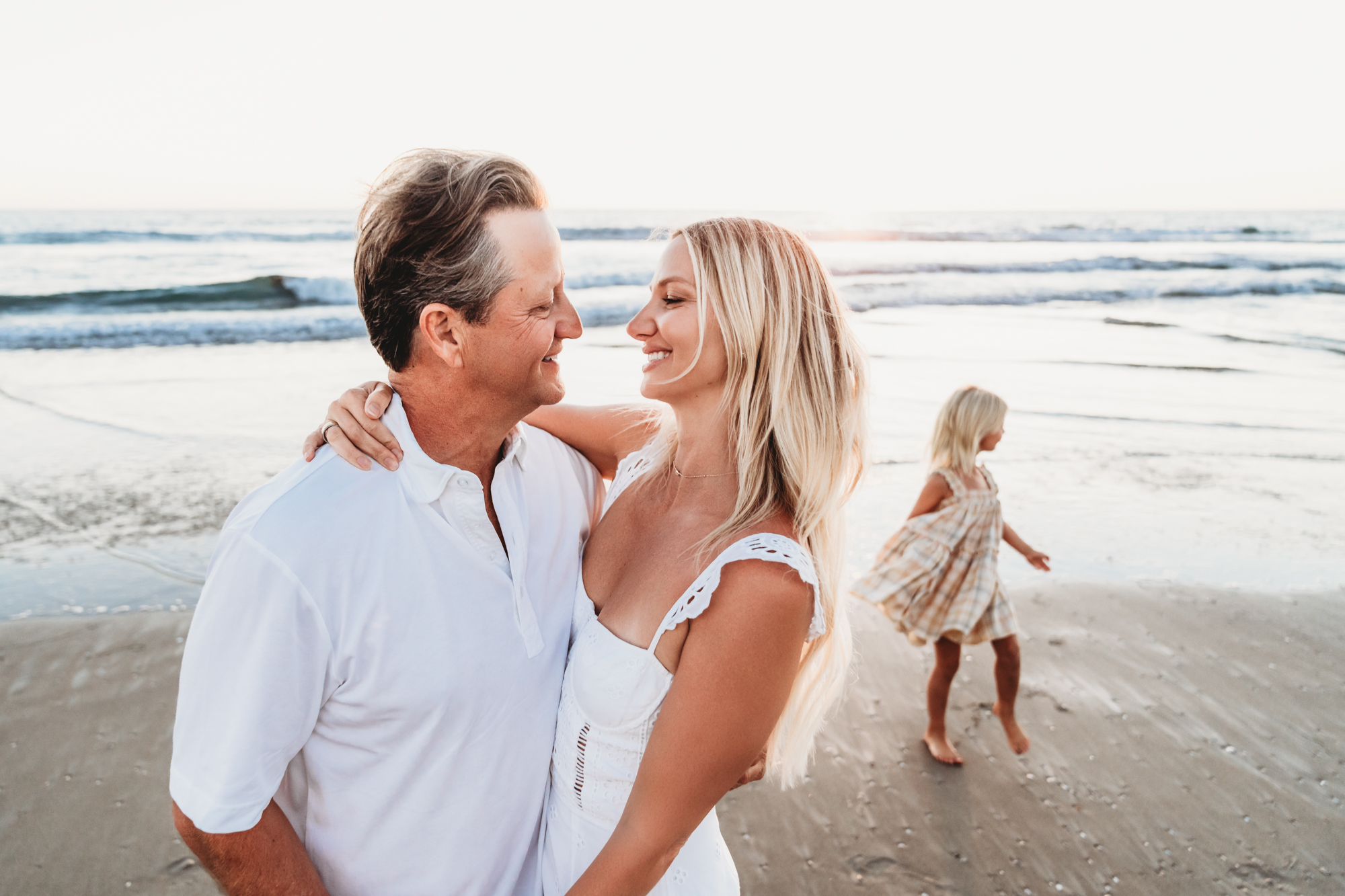 A man and wife embrace on Mission Beach while their young daughters play in the surf behind them. 