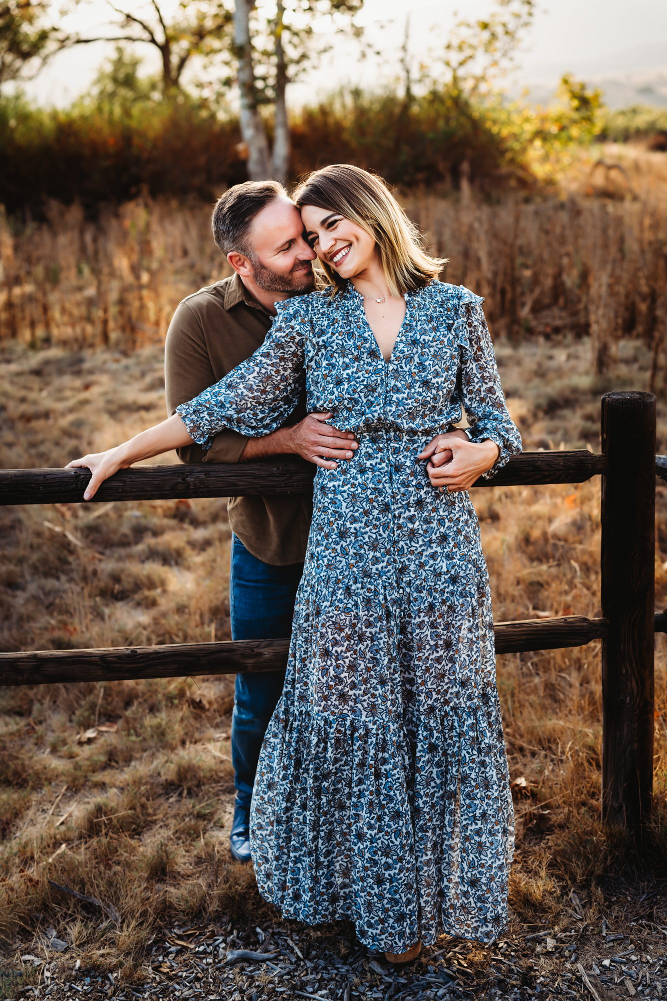 A man and wife lean against a wooden fence and embrace in Mission Trails Regional Park during a San Diego fall family photo session by Love Michelle Photography. 