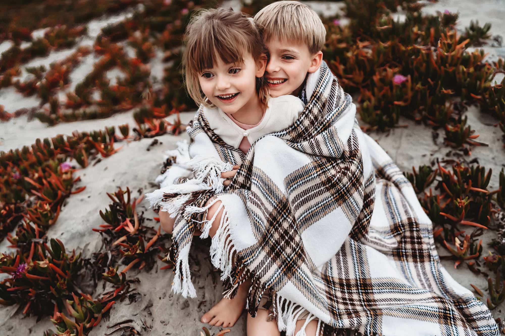 A blond boy and his little sister cuddle up together in a blanket while sitting on a Coronado Beach sand dune. 