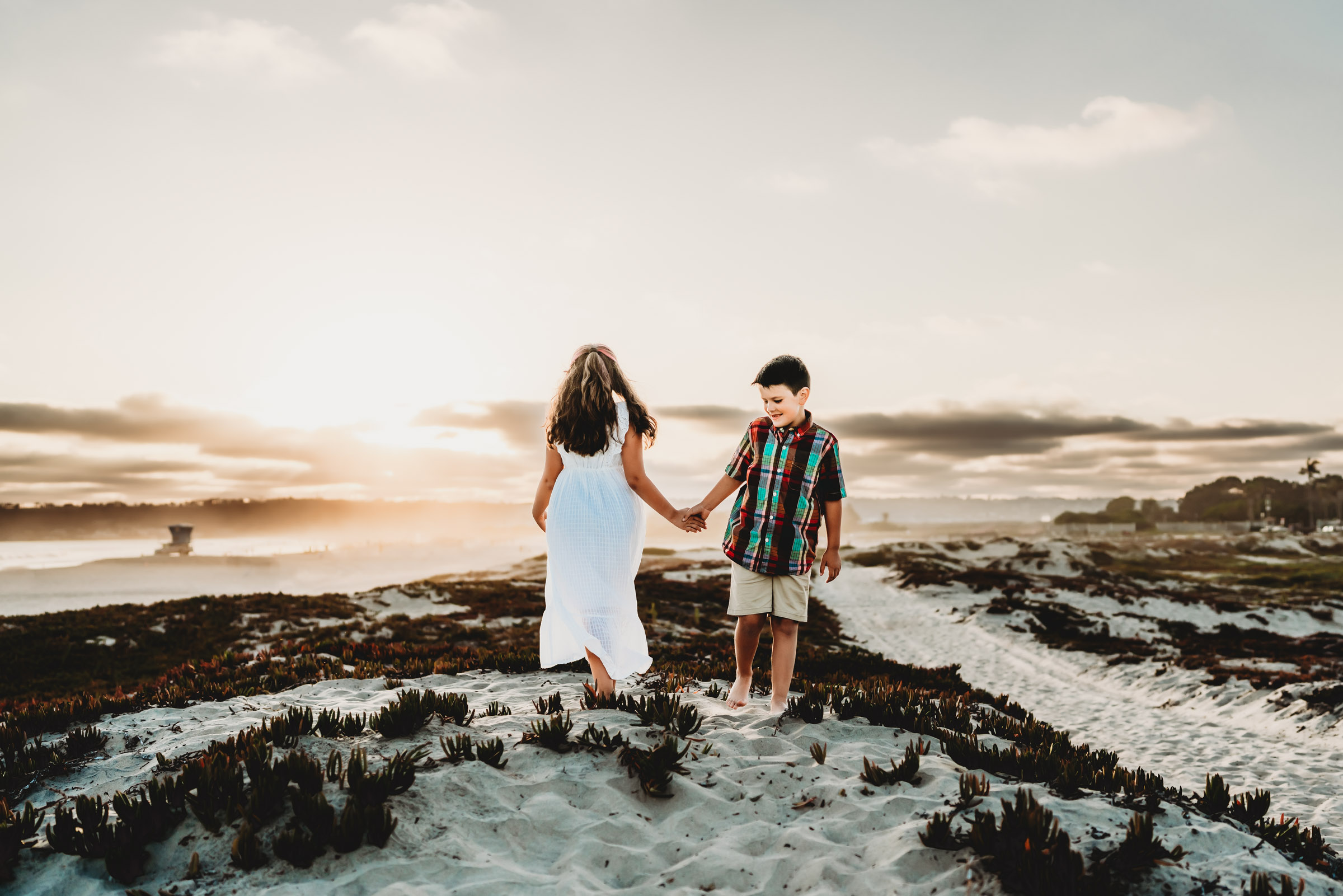 A boy and girl holding hands on a Coronado Beach sand dune during a lifestyle family beach photography session by Love Michelle Photography.