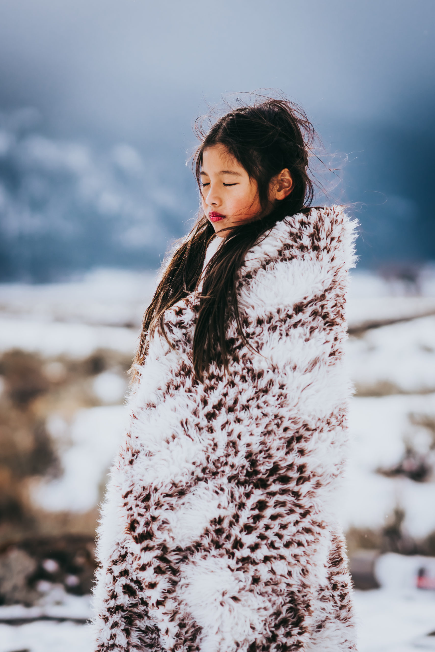 A young girl with long dark hair stands on a snowy field in Yellowstone National Park's Lamar Valley, wrapped in a furry blanket with her eyes closed.