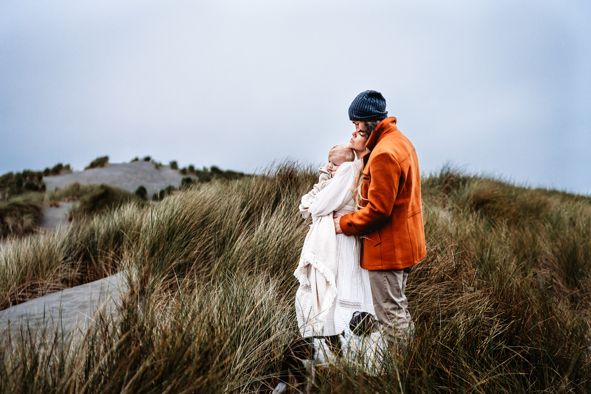 A man and woman stand on a grassy Oregon sand dune and embrace, while the woman is holding a baby.
