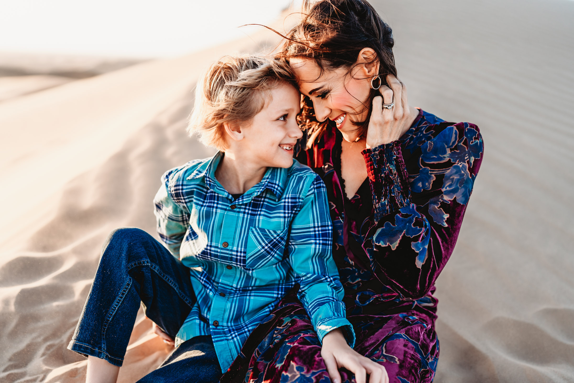 A woman and her son looking at each other and smiling while sitting on a sand dune during a lifestyle family photography session at the Imperial Sand Dunes in Glamis, California. 