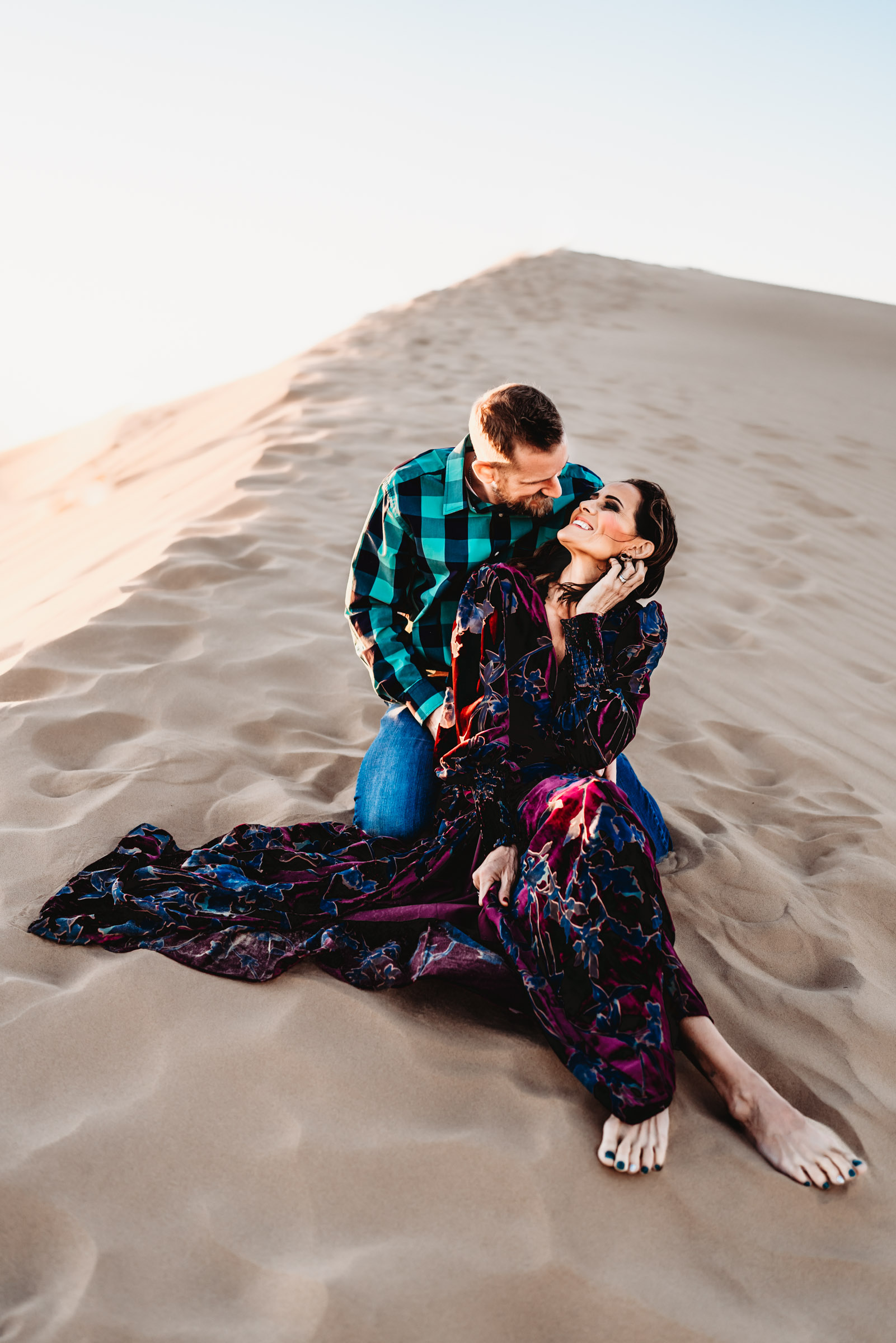 A man and woman sitting on a sand dune looking at each other during a lifestyle family photography session at the Imperial Sand Dunes in Glamis, California. 