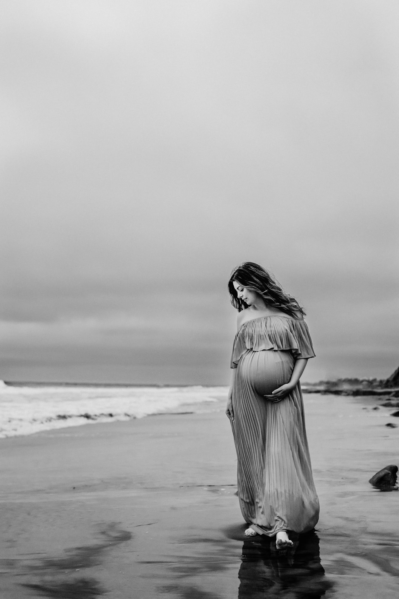 A pregnant woman walks on the beach in Del Mar with her hand on her belly and looks down at the sand. This is during a maternity photoshoot by San Diego's Love Michelle Photography