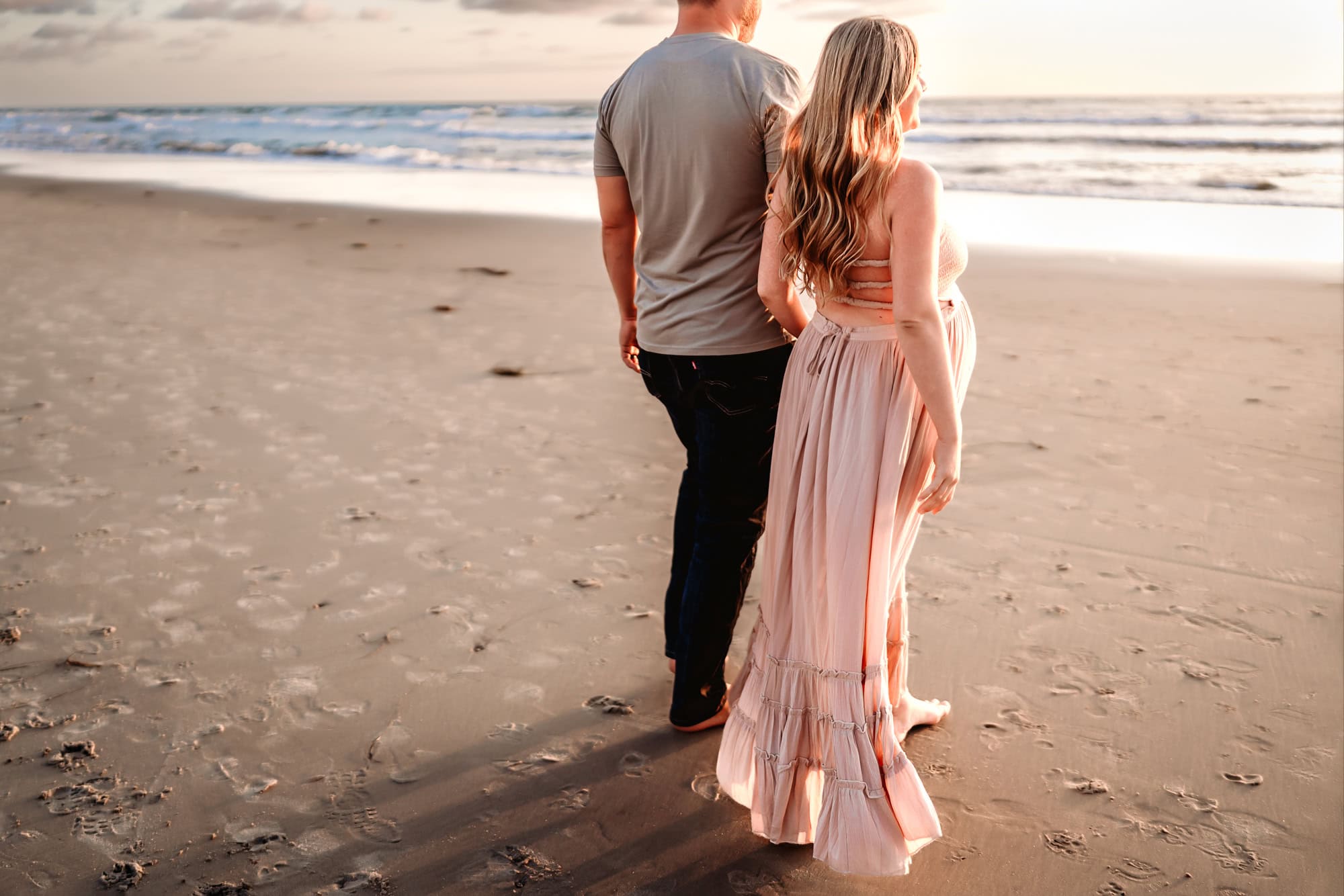 A pregnant woman wearing a long pink dress holds her husband's hand while they walk toward the shoreline in Del Mar, CA, during a maternity photoshoot by Love Michelle Photography. 
