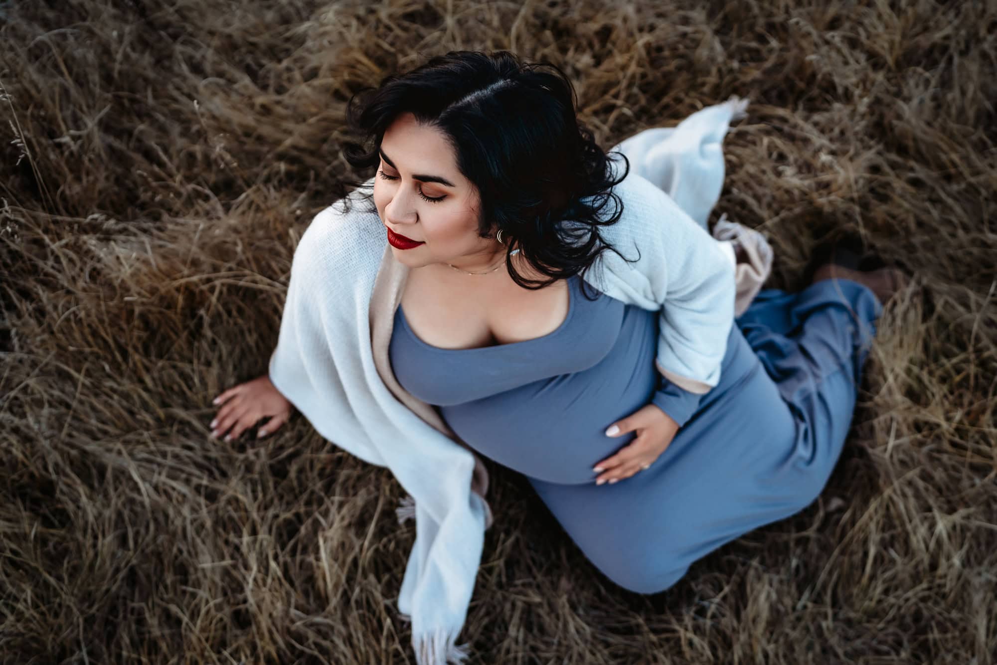 A pregnant woman in a blue dress with a white shawl sits in the grass, holding her belly, closing her eyes, and facing toward the sky. 