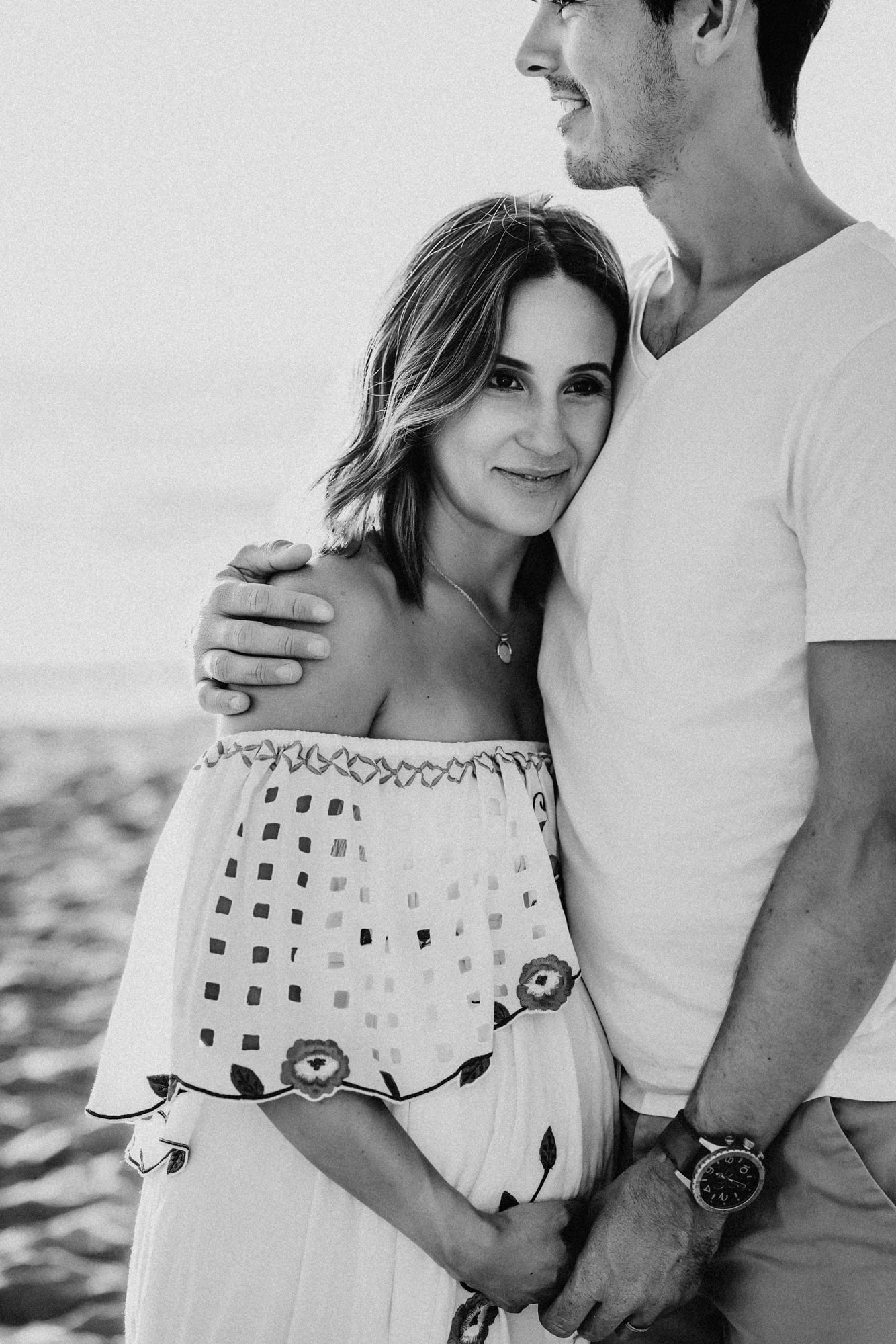 A pregnant woman wearing a bohemian white dress stands on a La Jolla beach while her husband embraces her during a maternity photoshoot by San Diego photographer Love Michelle Photography.