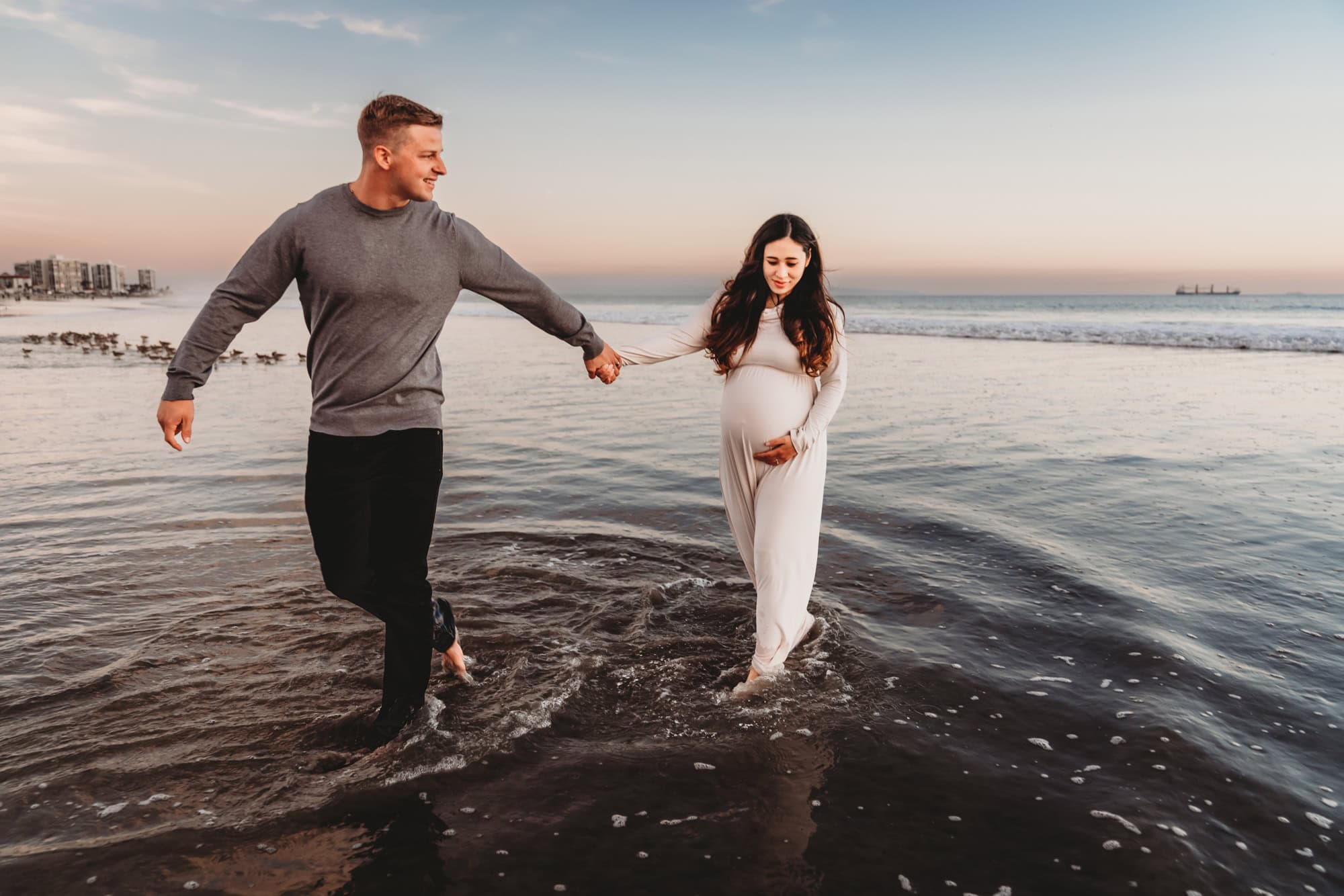 A man on Coronado Beach holds his pregnant wife's hand and leads her through the shoreline during a maturity photoshoot in San Diego, CA. 