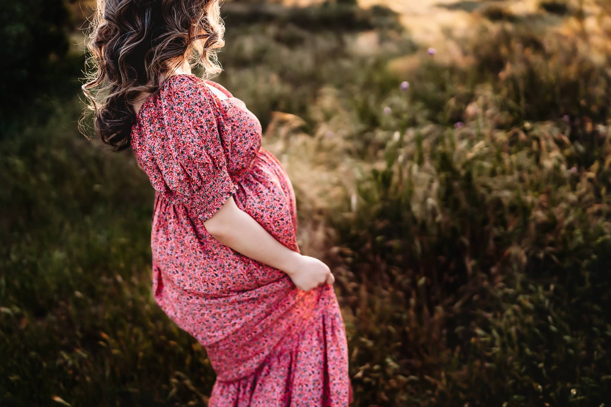 A pregnant woman in a pink dress holds her skirt while walking through green field during a maternity photo session by Love Michelle Photography in San Diego, CA.