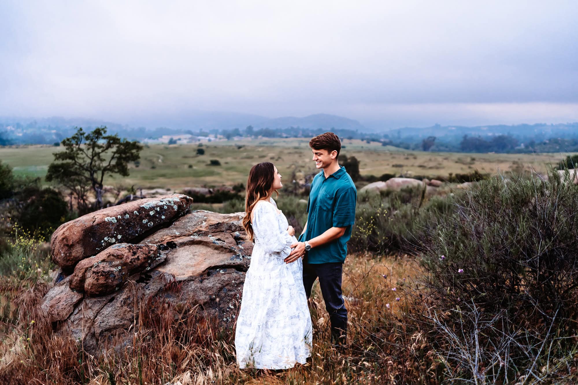 A pregnant woman and her husband hold hands and gaze at each other while standing on a field next to large boulders in San Diego, CA. 