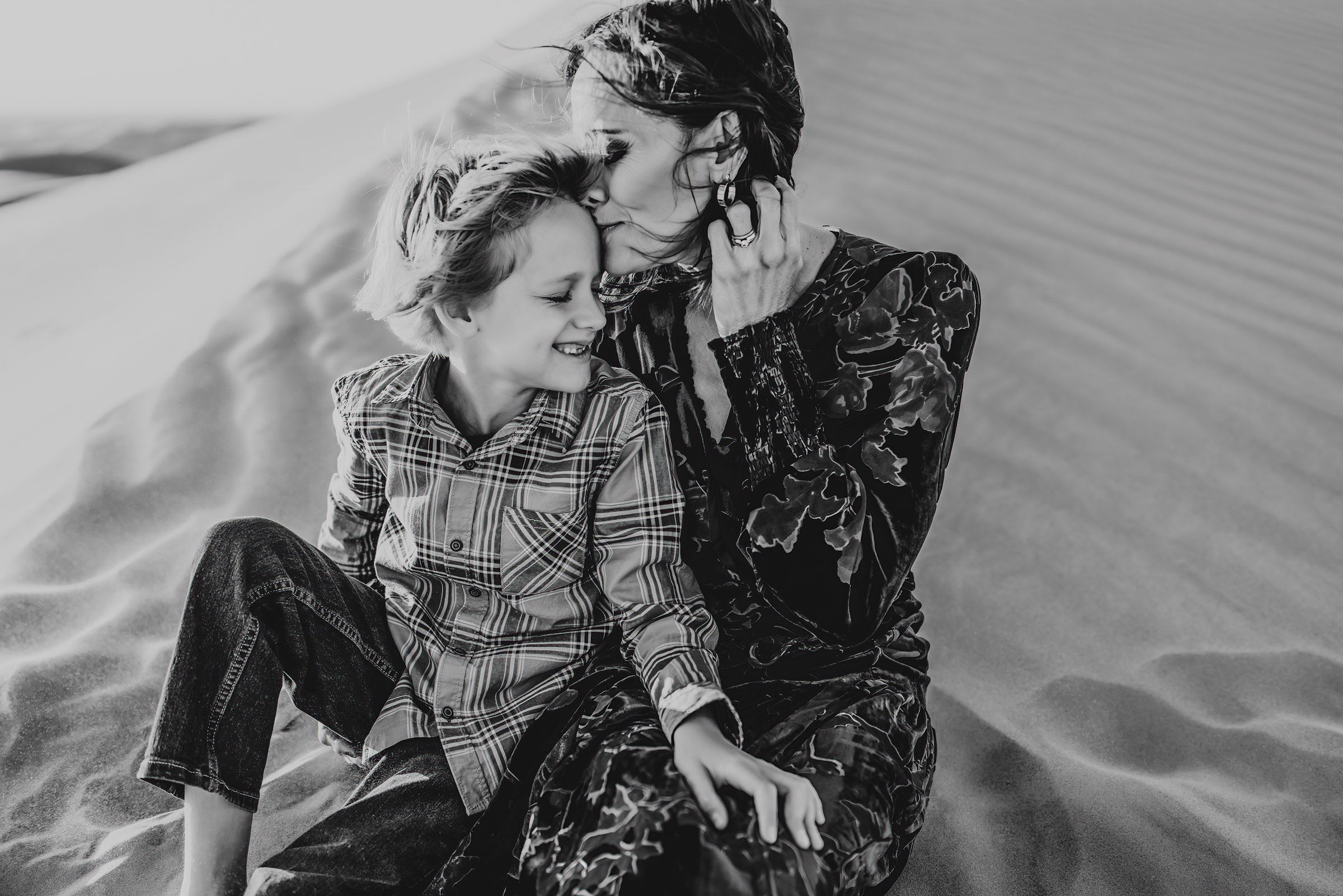 A woman kissing her son while sitting on a sand dune during a lifestyle family photography session at the Imperial Sand Dunes in Glamis, California. 