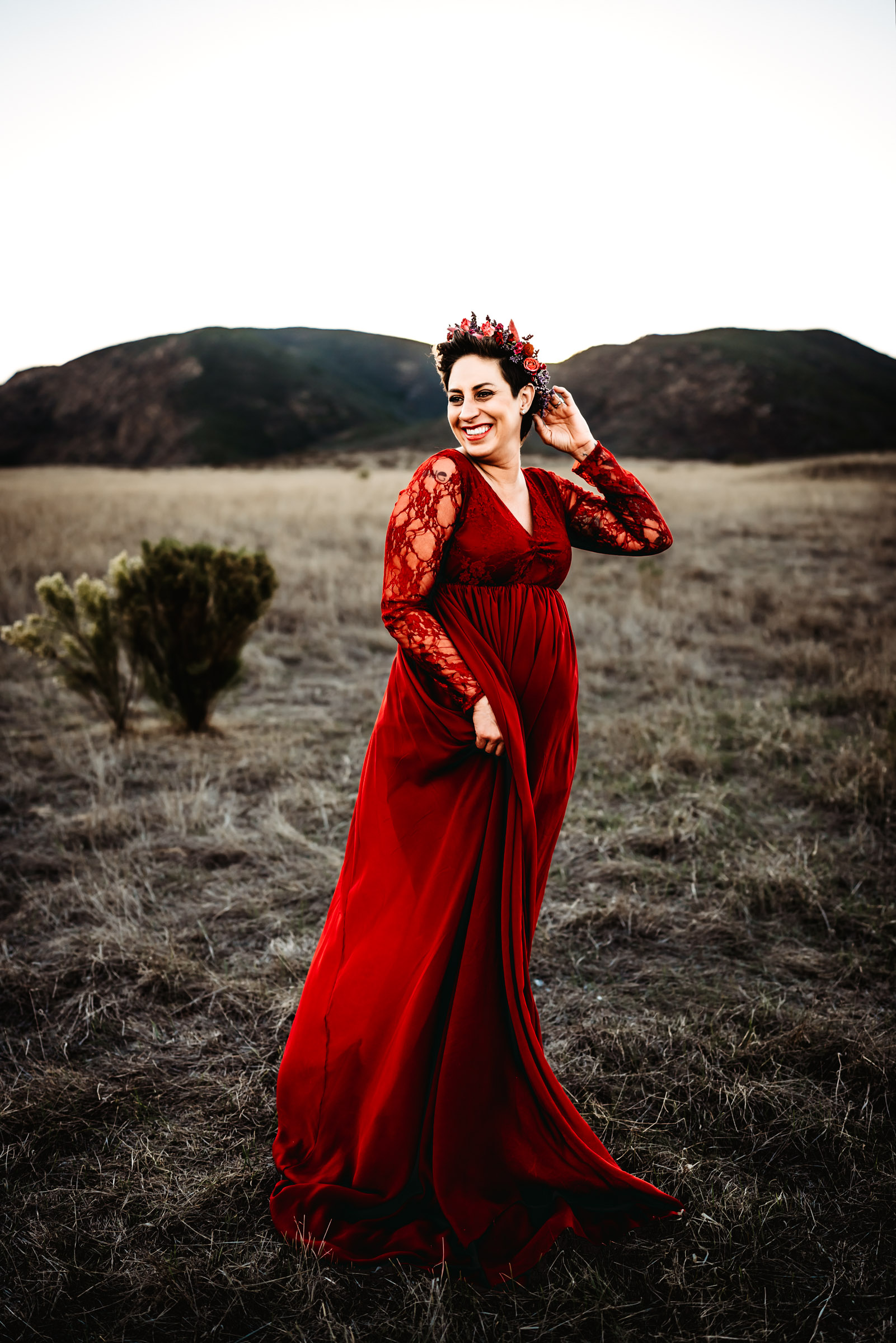 Pregnant woman wearing a red dress and flower crown laughing during a San Diego lifestyle maternity session in Mission Trails Regional Park