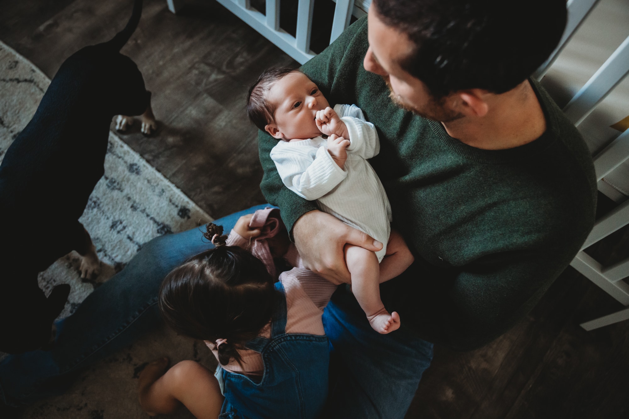 Dad sitting on floor next to crib, holding his newborn baby during a San Diego lifestyle newborn photography session