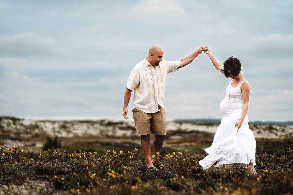 A pregnant woman and her husband holding hands in the wildflowers on Coronado beach during a San Diego lifestyle maternity photo session by Love Michelle Photography.