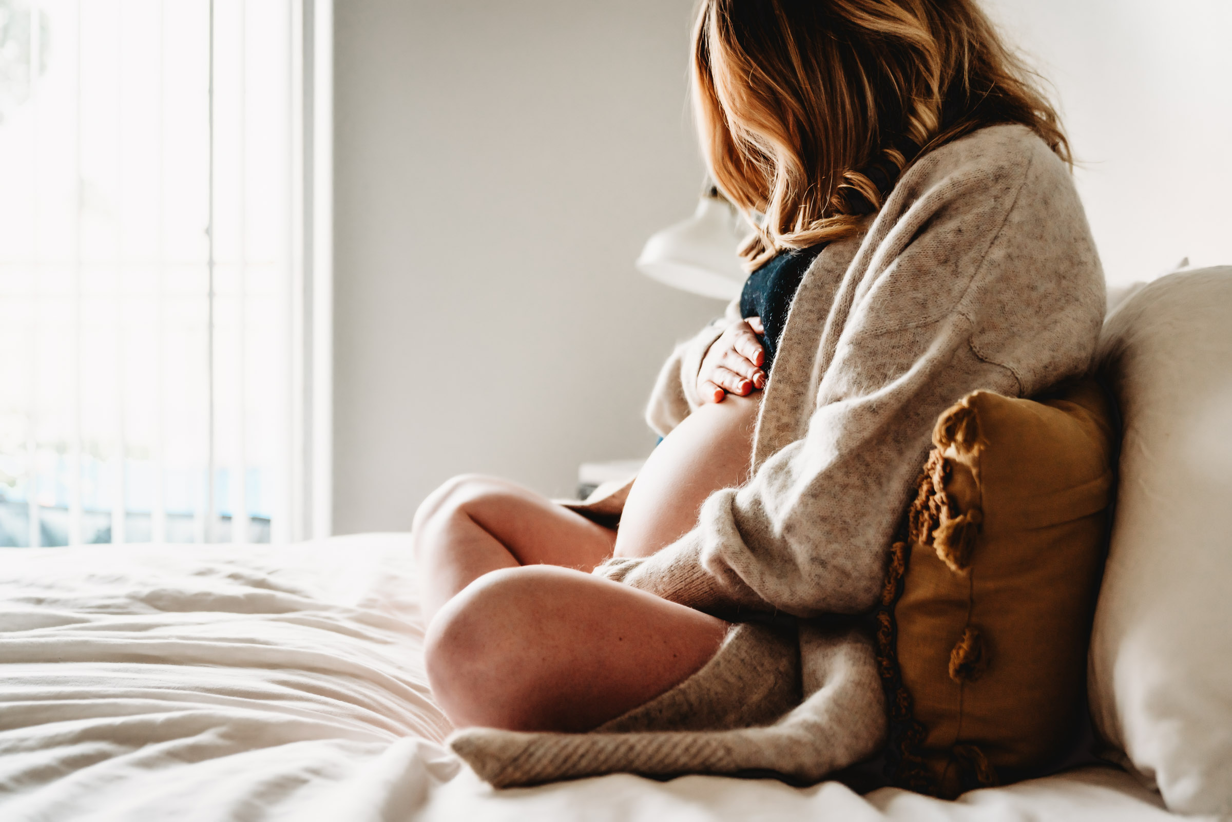 A pregnant woman sitting on her bed looking at and holding her belly during a San Diego lifestyle in-home maternity session by Love Michelle Photography.
