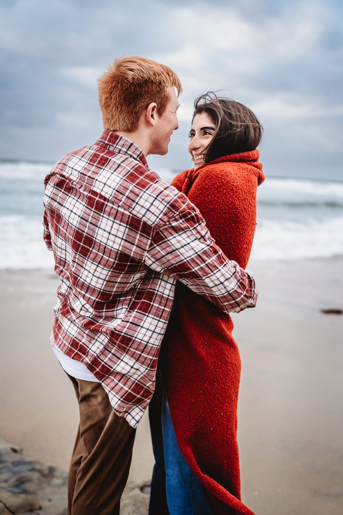 A young couple wrapped up in a warm blanket smiling at each other on a San Diego beach during a San Diego beach engagement photo session by Love Michelle Photography.