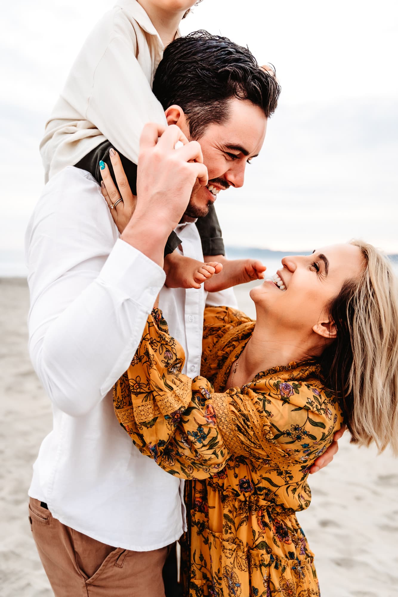 Man smiling and holding his baby boy on his shoulders while looking down at his wife during a Coronado Beach family photo session by Love Michelle Photography.