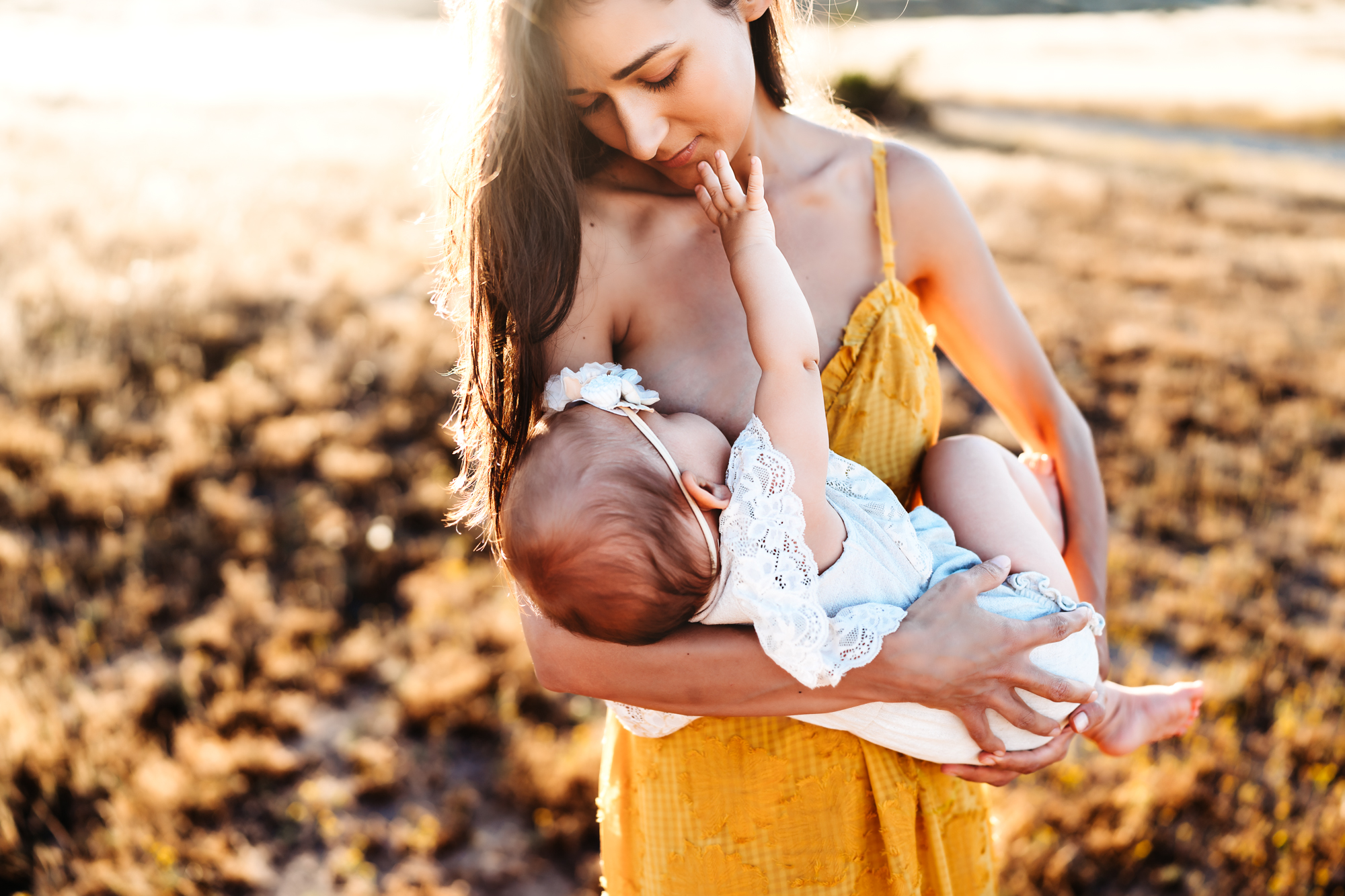 Woman wearing a yellow dress standing in a field in Mission Trails Regional Park breastfeeding her baby girl during a lifestyle family photo session by Love Michelle Photography.