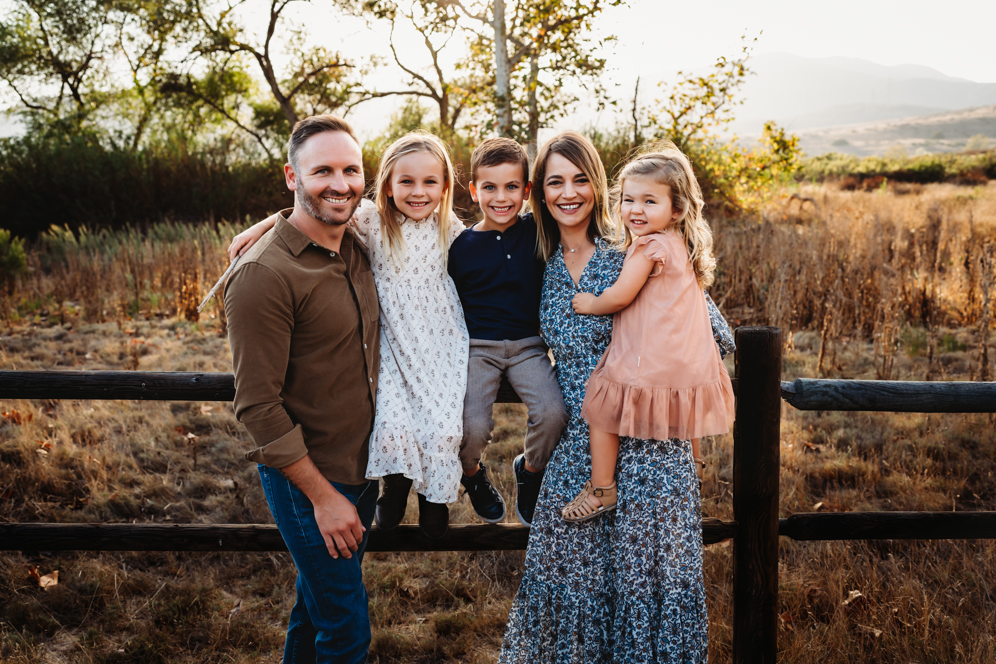A family of 5 leaning against a wooden fence in Mission Trails Regional Park while smiling at the camera during a family photo session. 