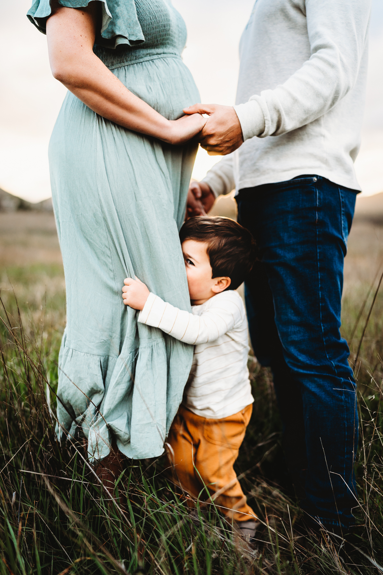 Baby boy holding on to mom's skirt playing peekaboo on a green field in Mission Trails Regional Park doing a lifestyle maternity photo session by Love Michelle Photography. 