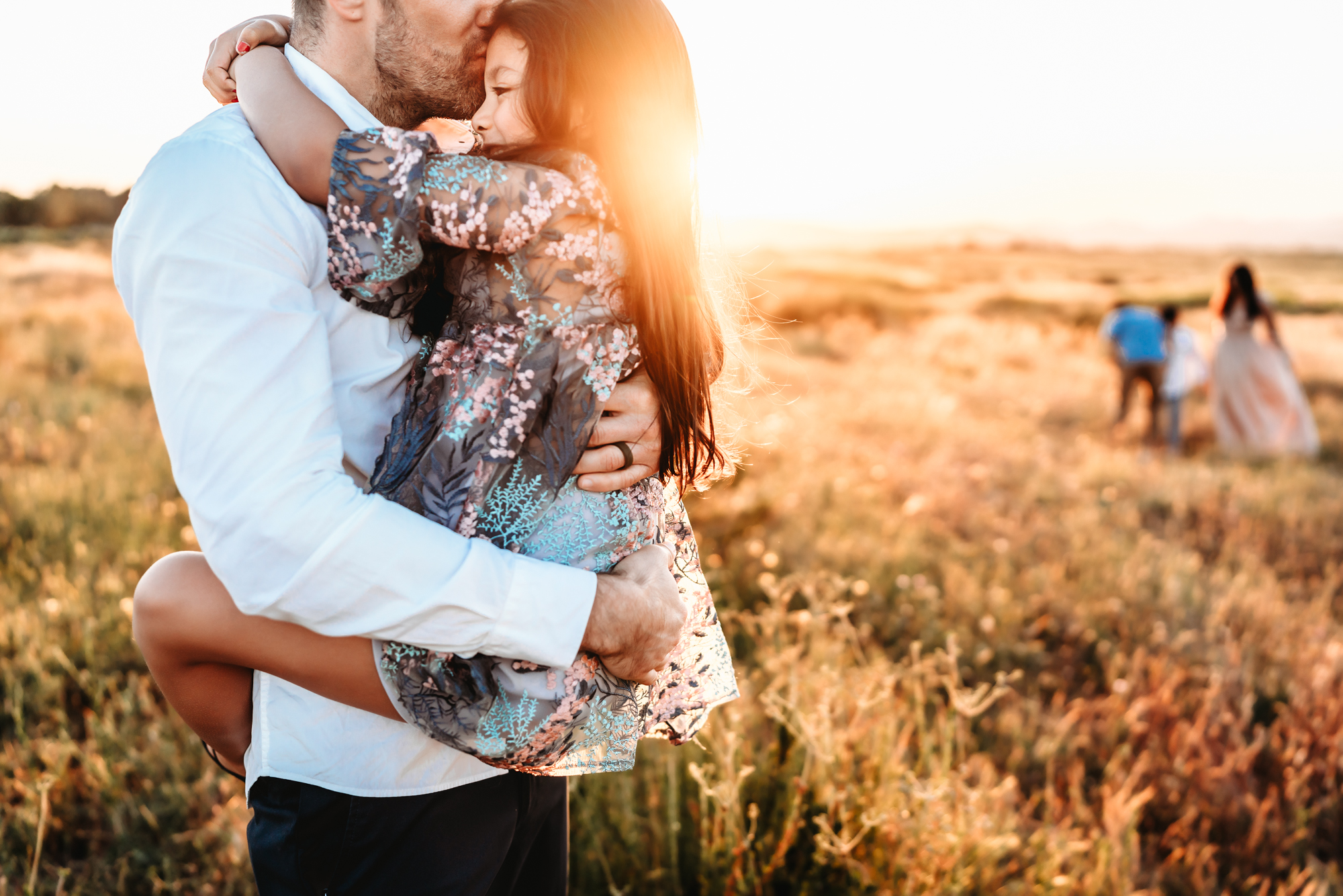 A man wearing a white dress shirt stands in a golden field at sunset, kissing his young daughter on the forehead while he holds her. The rest of the family is walking in the background.