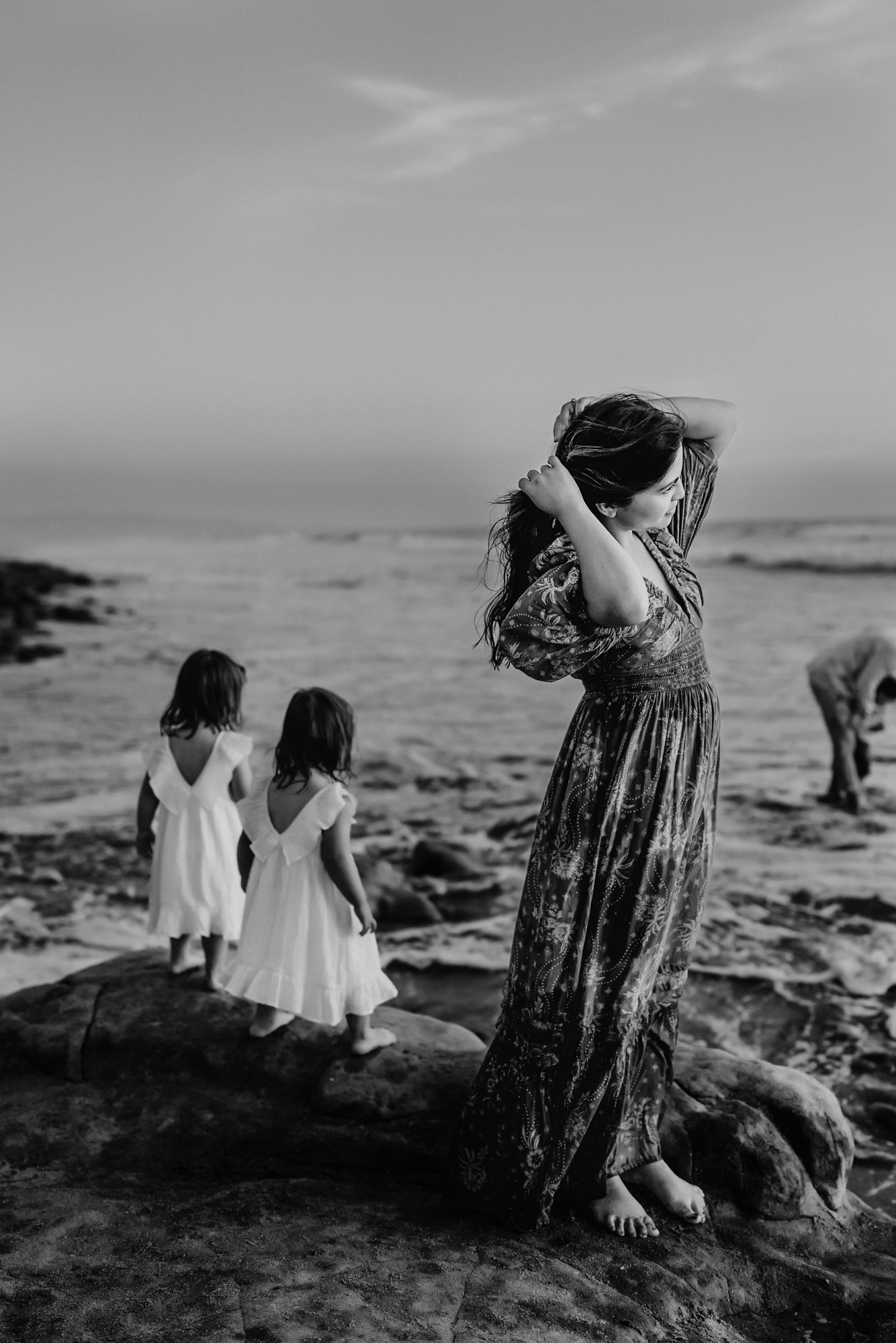 A woman standing on a rock fixing her hair, with her twin daughters behind her, on the beach in Del Mar, California.
