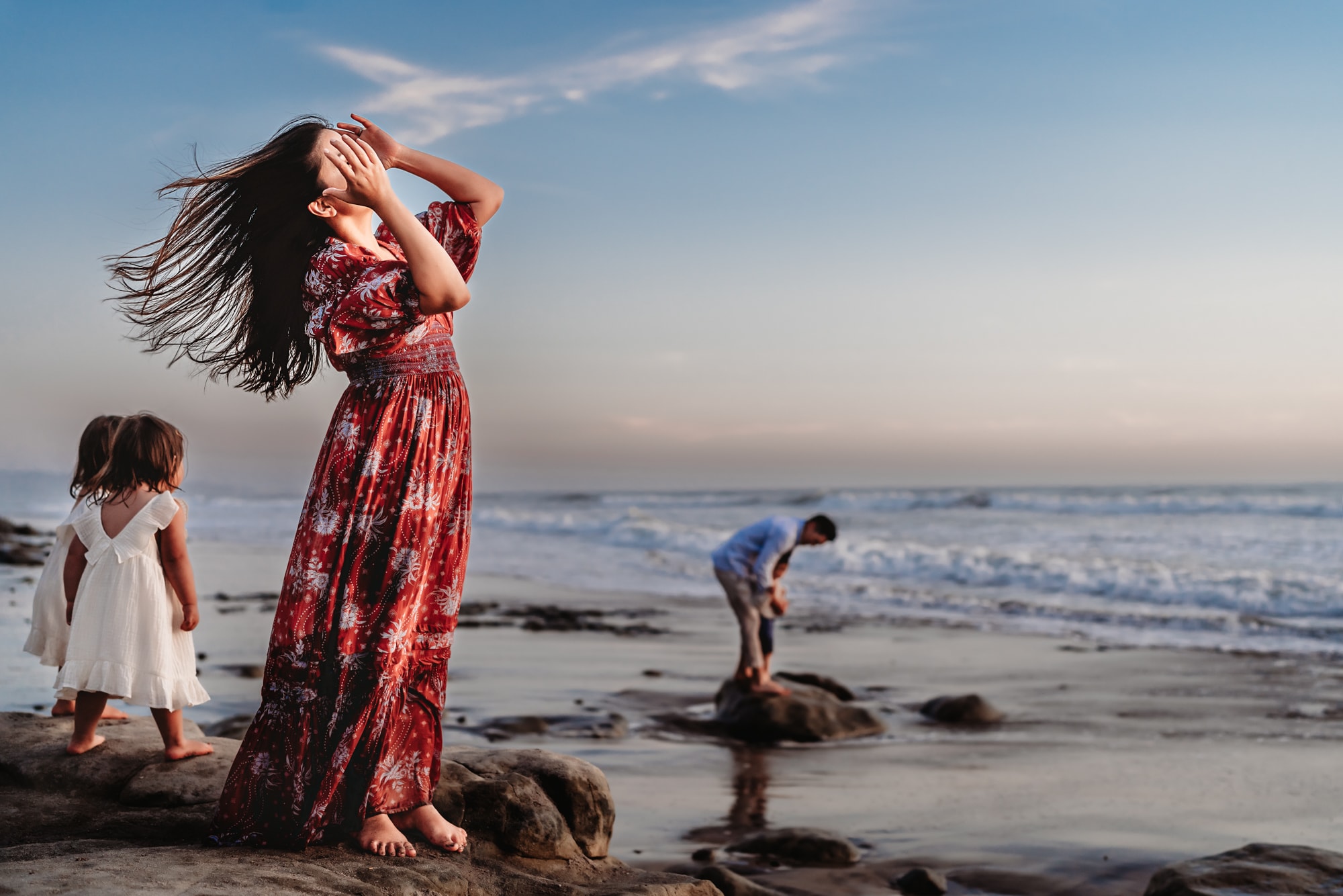A woman in a red dress standing on a rock on a beach in Del Mar, while her family plays around her. 