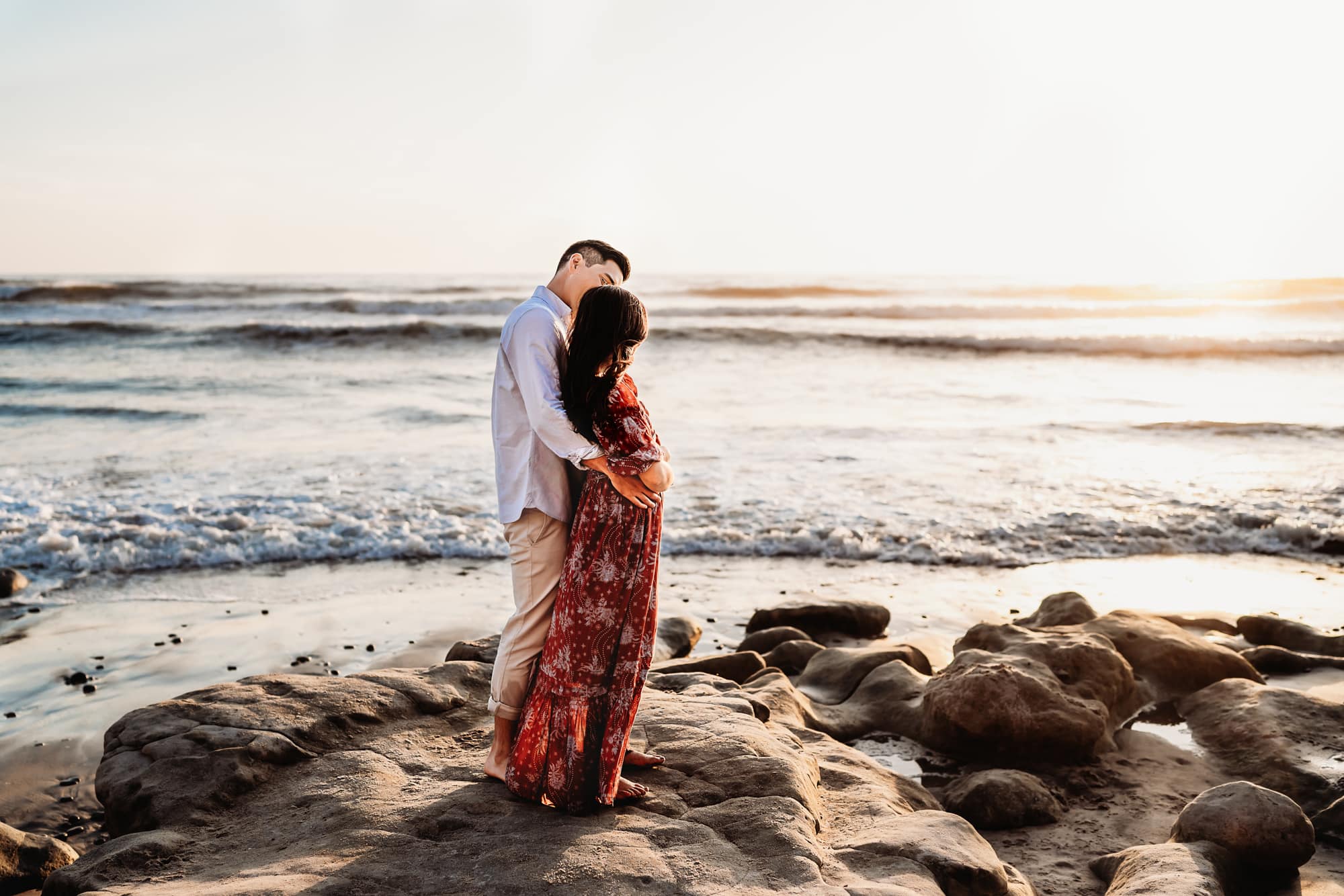 A husband and wife facing away from the camera, standing on a rock on a beach in Del Mar, California. 