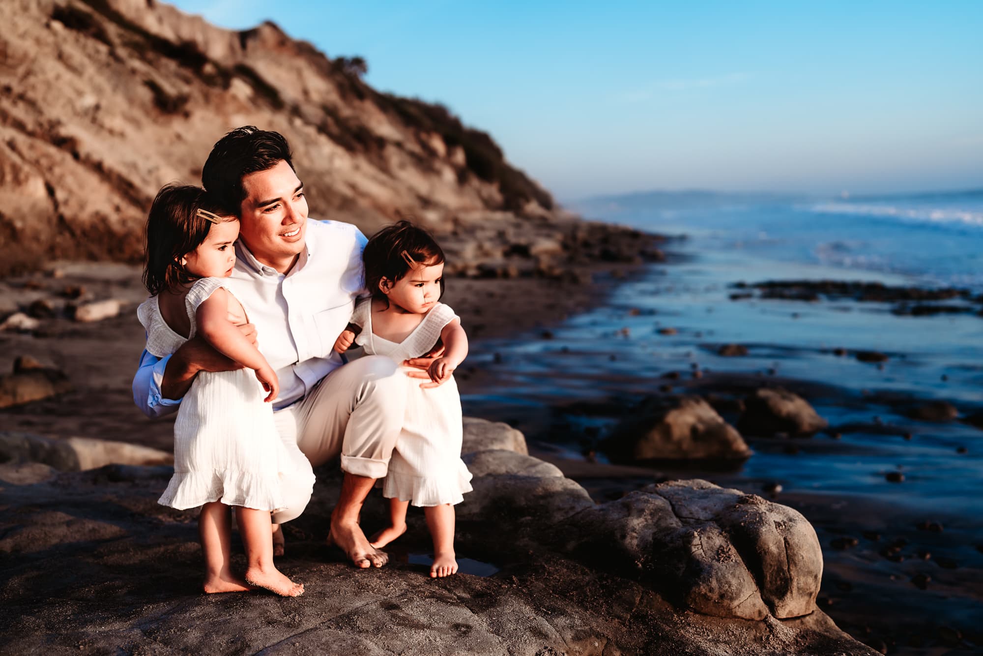 A father crouching next to his twin 2 year old daughters, looking at the water during a lifestyle family photography session on a beach in Del Mar, California. 