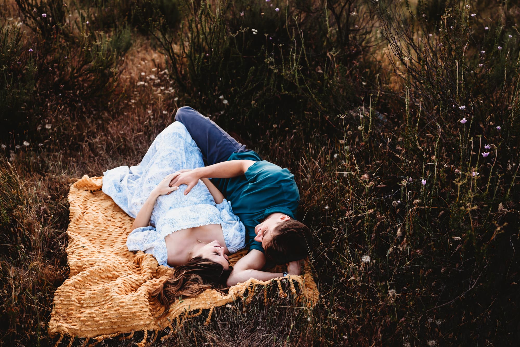 A pregnant woman and her husband lying on a yellow blanket in a field during a San Diego lifestyle maternity photography session.