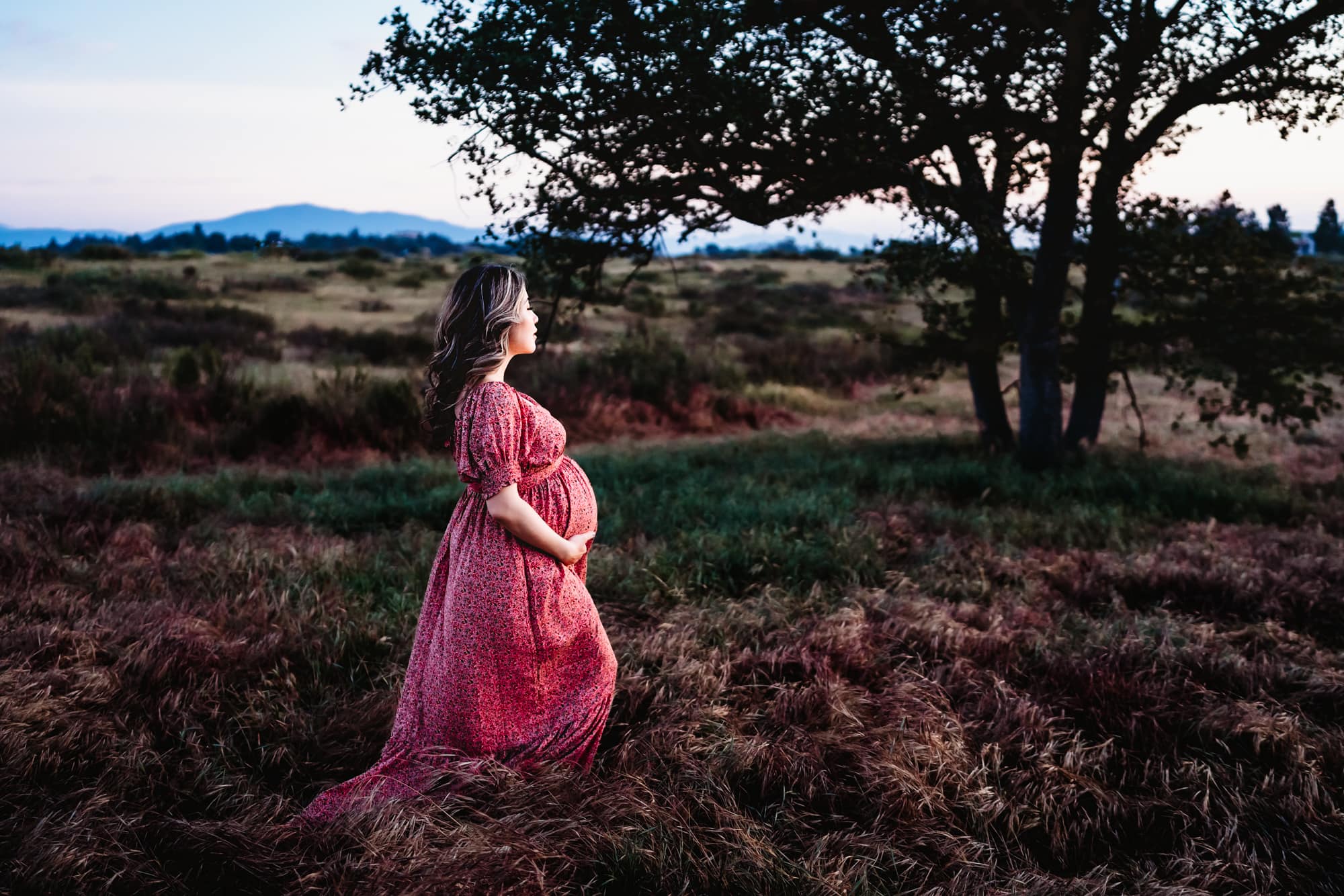 A pregnant woman in a pink dress walking through a field of tall grass at sunset during a San Diego lifestyle maternity session by Love Michelle Photography.