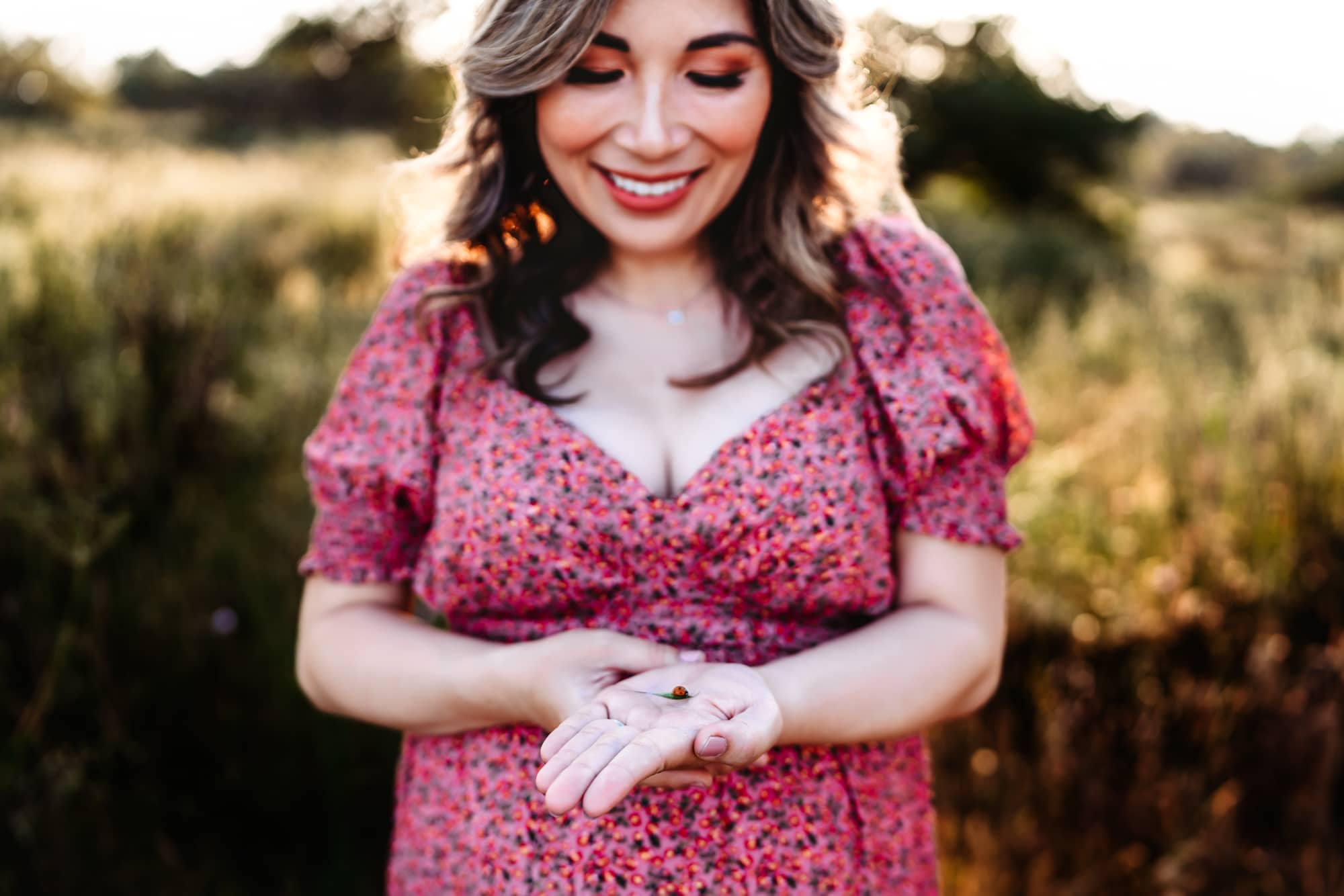 A pregnant woman in a pink dress looking down and smiling at a ladybug in her hand during a San Diego lifestyle maternity session by Love Michelle Photography.
