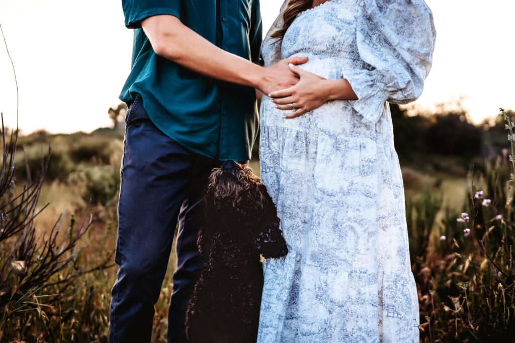 A man puts his hand on his pregnant wife's belly while their black dog looks up at them during a San Diego maternity photo session.