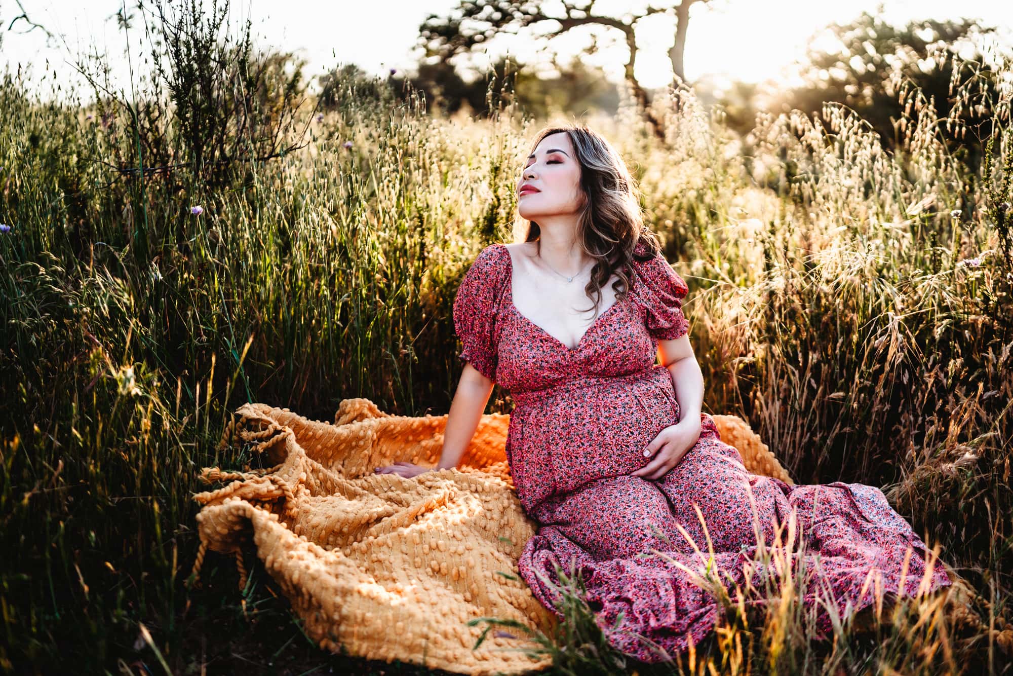 A pregnant woman in a pink dress sitting on a yellow blanket holding her belly and closing her eyes during a San Diego lifestyle maternity session by Love Michelle Photography.