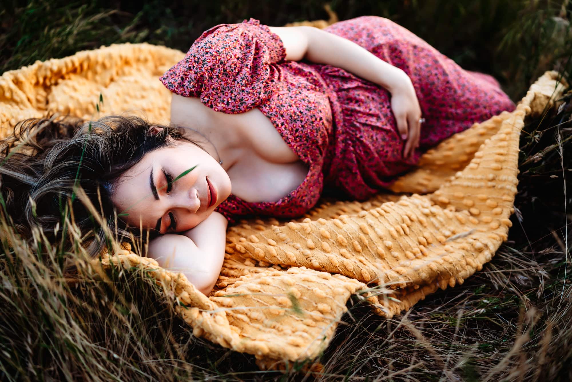 A pregnant woman in a pink dress laying on a yellow blanket holding her belly and closing her eyes during a San Diego lifestyle maternity session by Love Michelle Photography.