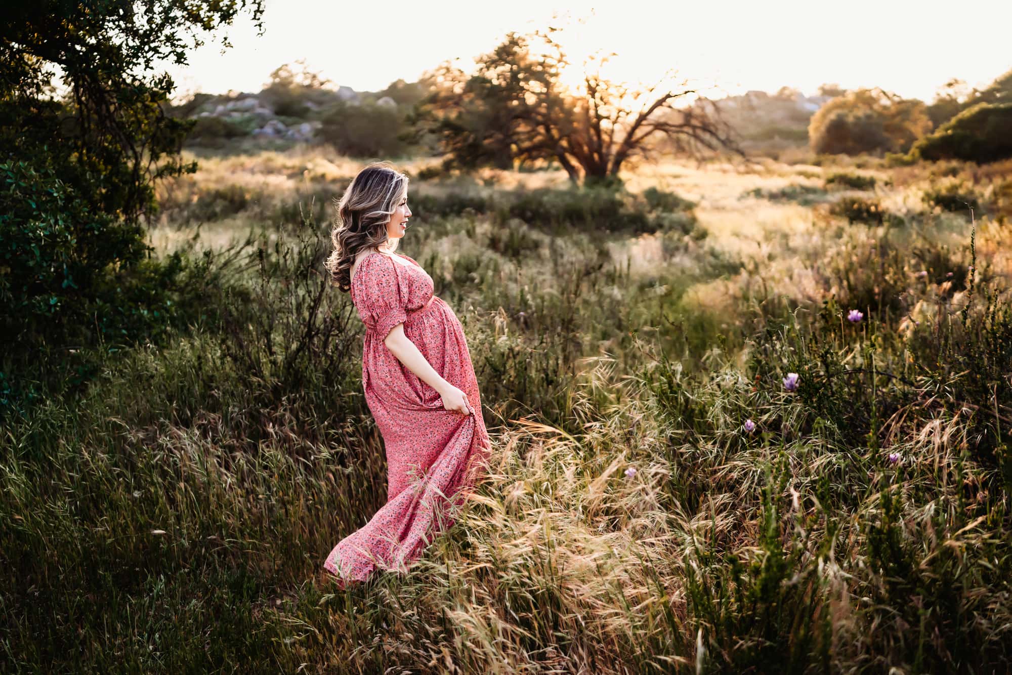 A pregnant woman in a pink dress walking through a field of tall grass at sunset during a San Diego lifestyle maternity session by Love Michelle Photography.
