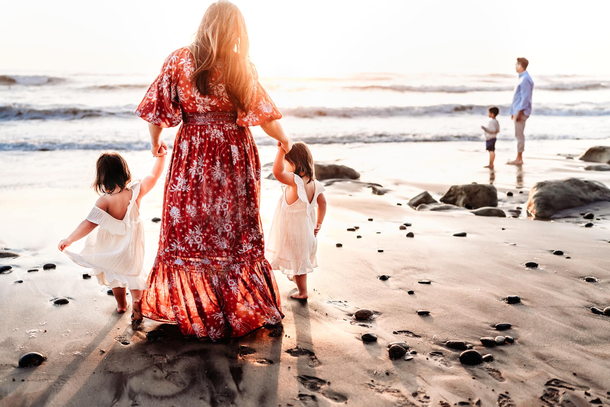 A woman in a long red dress holding her twin daughters' hands and walking on the sand on a Del Mar beach. 