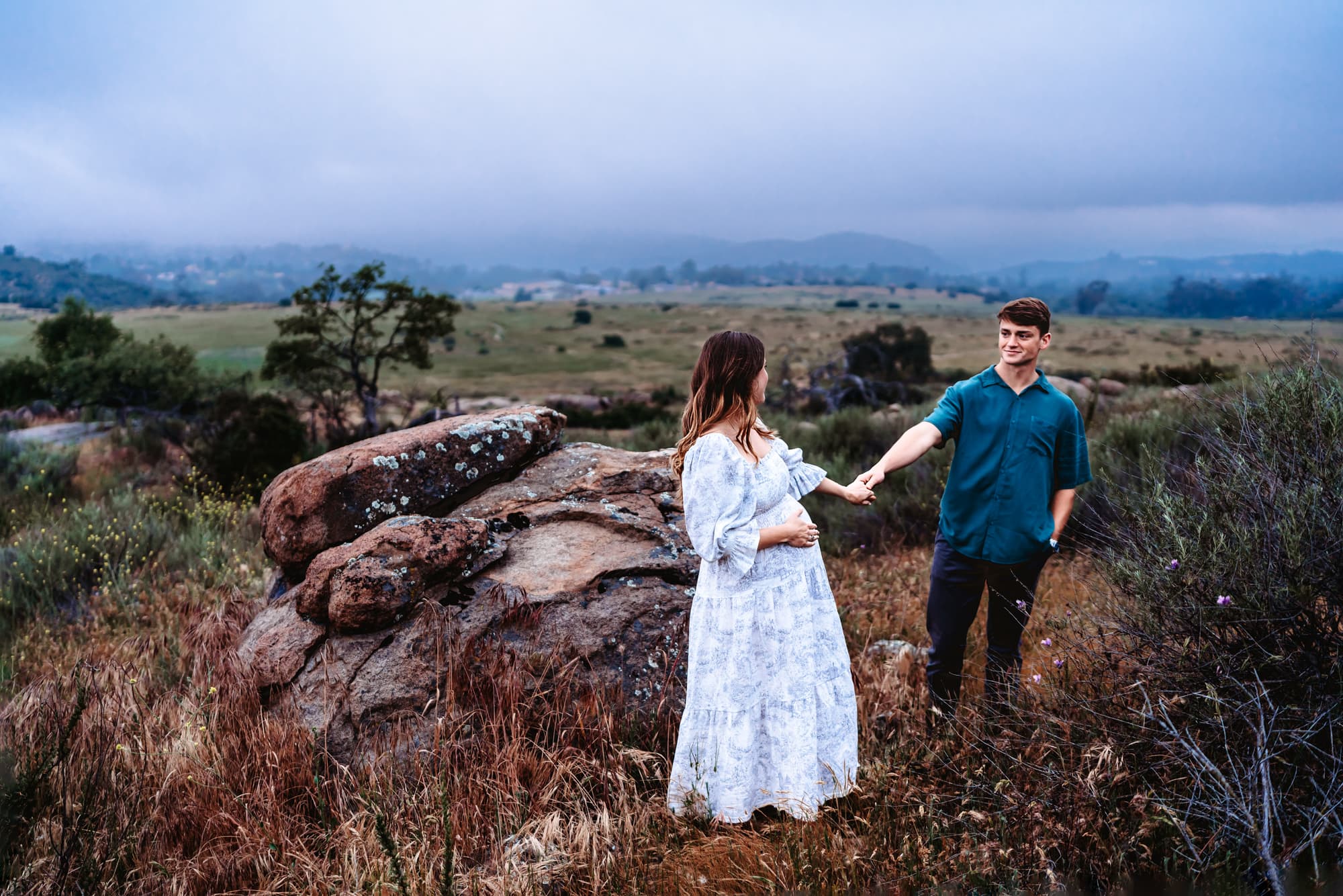 A husband and his pregnant wife hold hands and look at eachother in front of a giant rock during a lifestyle maternity photo session in Alpine, CA.