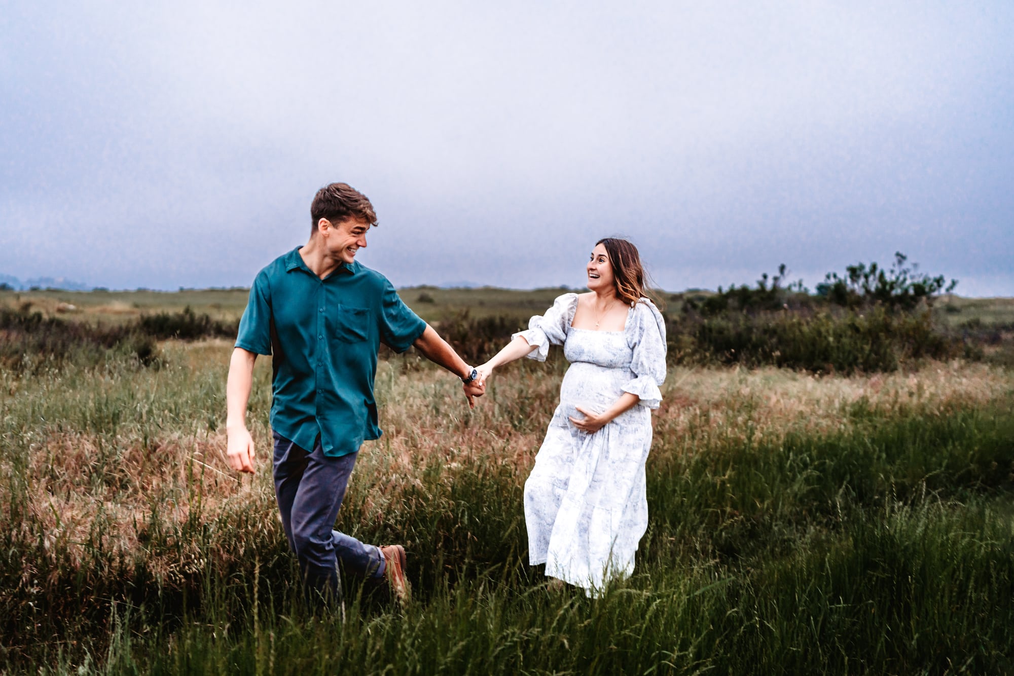 A husband holding his pregnant wife's hand and running while she holds her belly and laughs during a lifestyle maternity photo session in San Diego. 