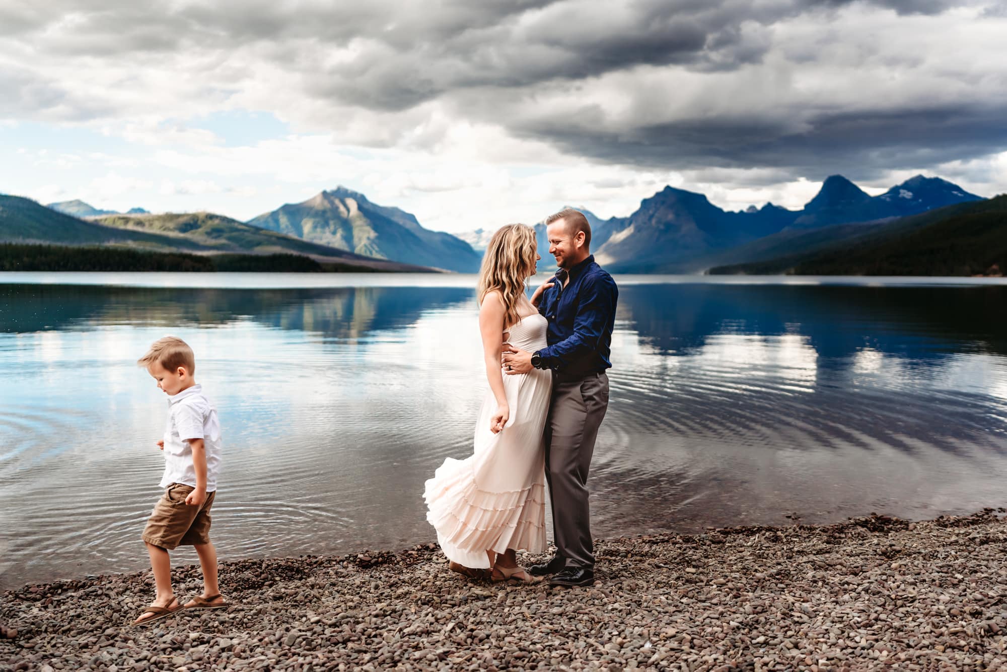 A man and woman embrace on the shores for Glacier National Park's Lake McDonald during a vacation family photography session by travel family photographer Love Michelle Photography. 
