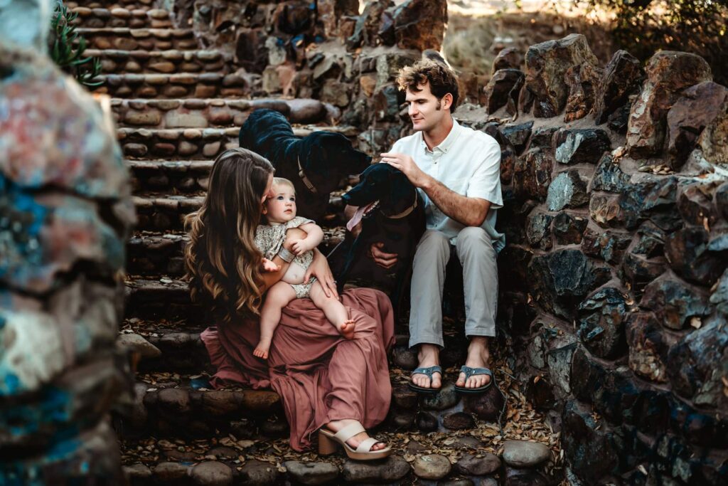 A couple and their baby sit on stone stairs with their dogs during a San Diego family photography session by Love Michelle Photography.