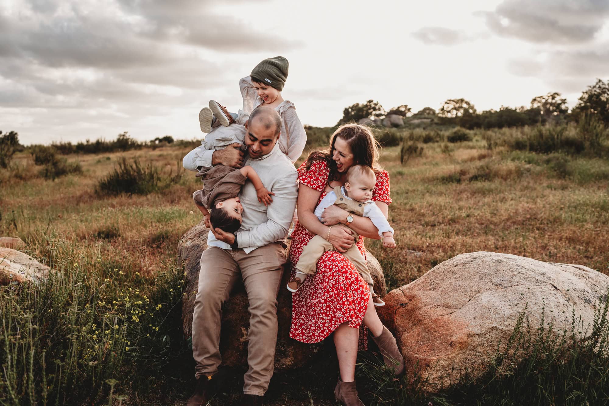 A family of 5 sits on a large boulder, laughing together while the kids act a little crazy. 