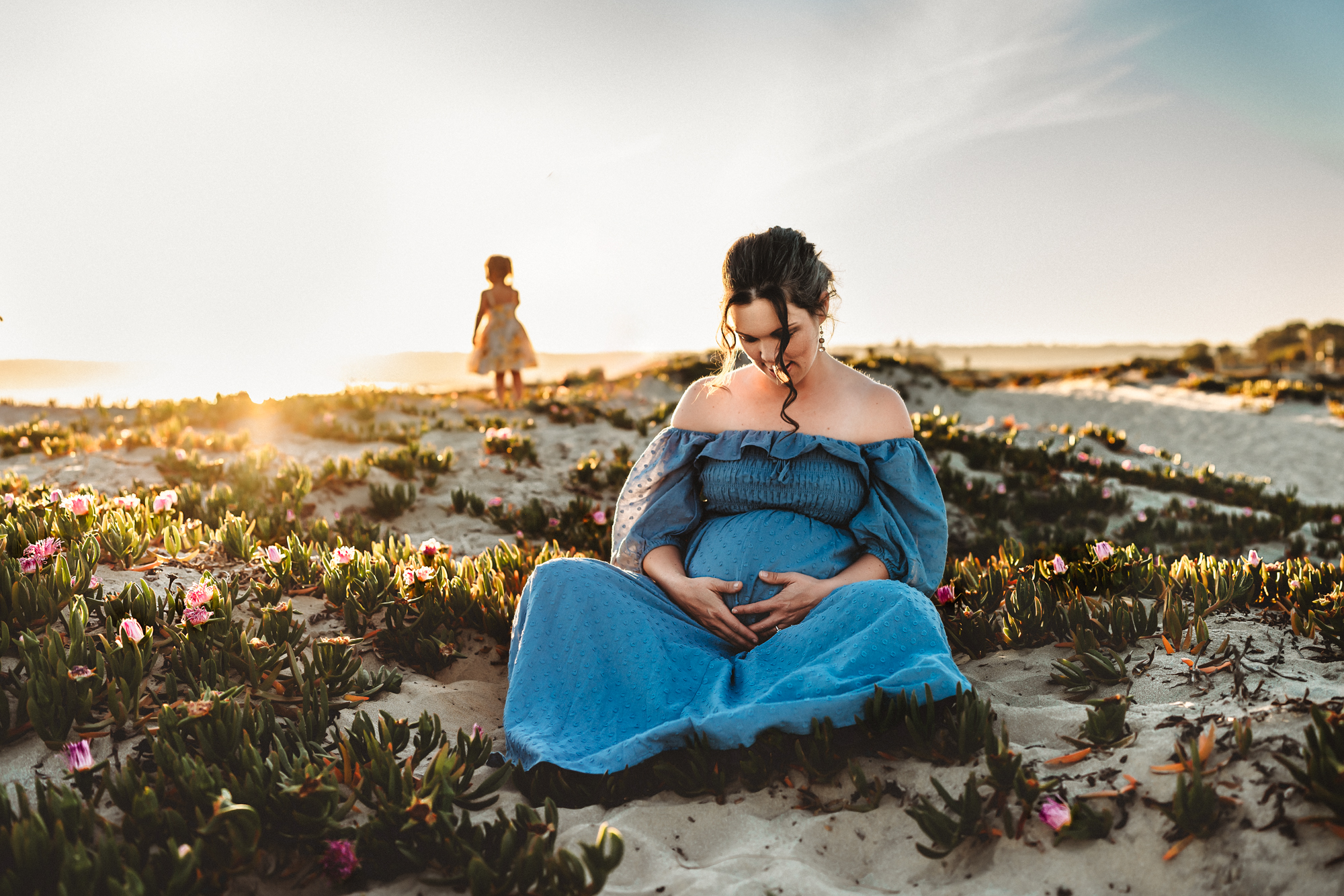 A pregnant woman in a long blue dress sits on a Coronado sand dune holding her belly. She's surrounded by greenery, and her toddler is behind her, looking at the sunset. This is during a San Diego maternity photography session by Love Michelle Photography 