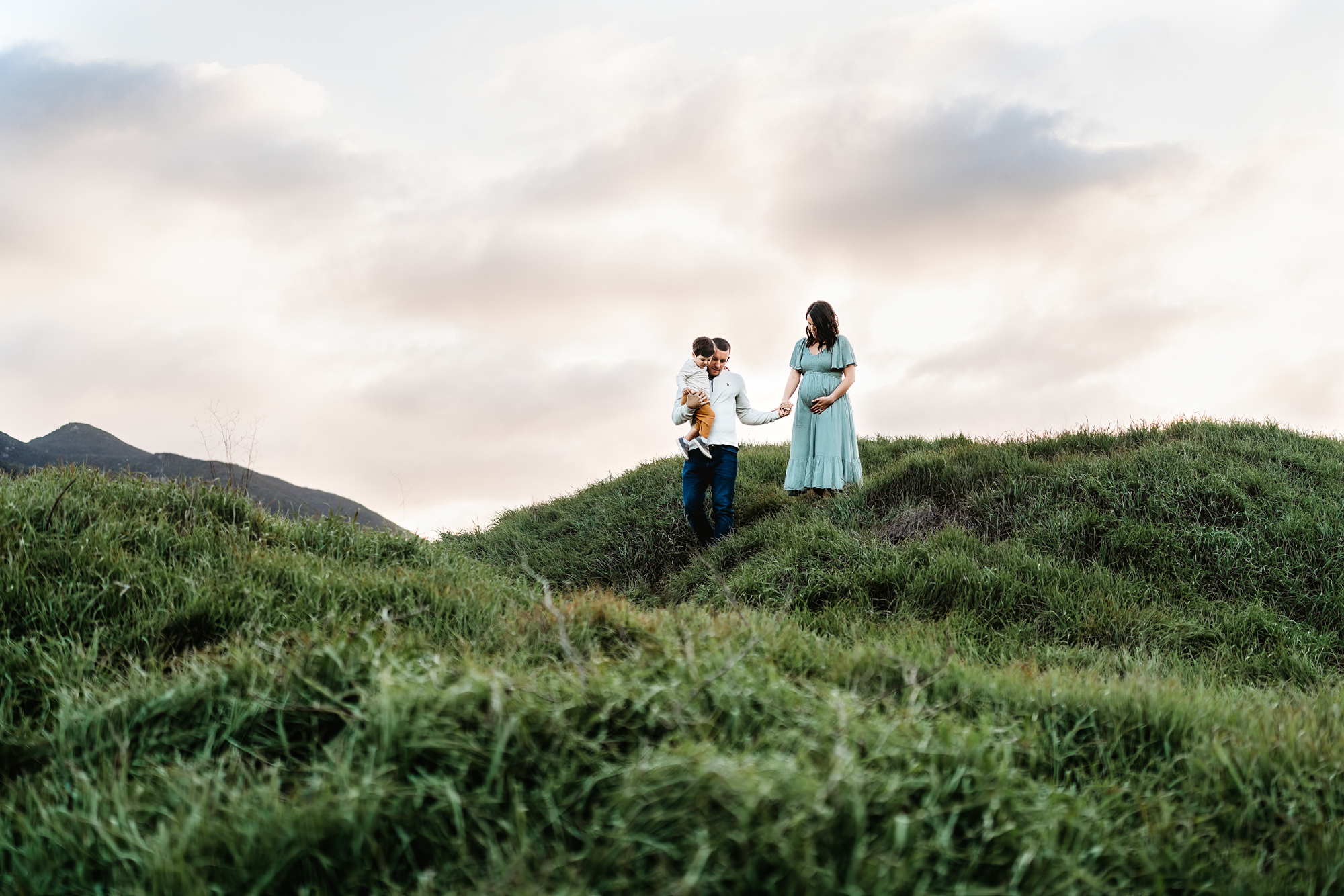 A man holds his toddler son and his pregnant wife's hand while walking down a green hillside during a San Diego lifestyle maternity photo session by Love Michelle Photography.
