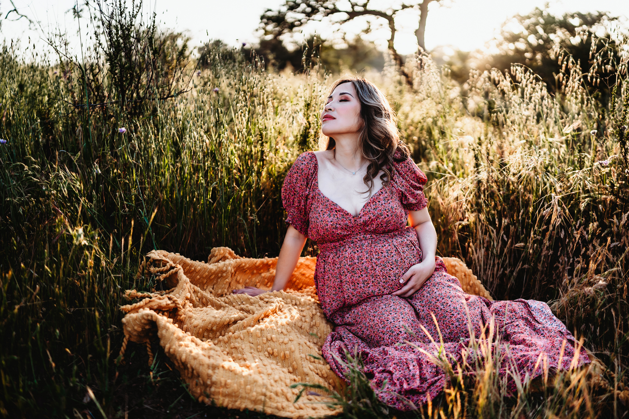A pregnant woman wearing a pink dress and sitting on a yellow blanket in long grass holds her belly and lifts her face to the sun. This is during a San Diego lifestyle maternity photo session by Love Michelle Photography.