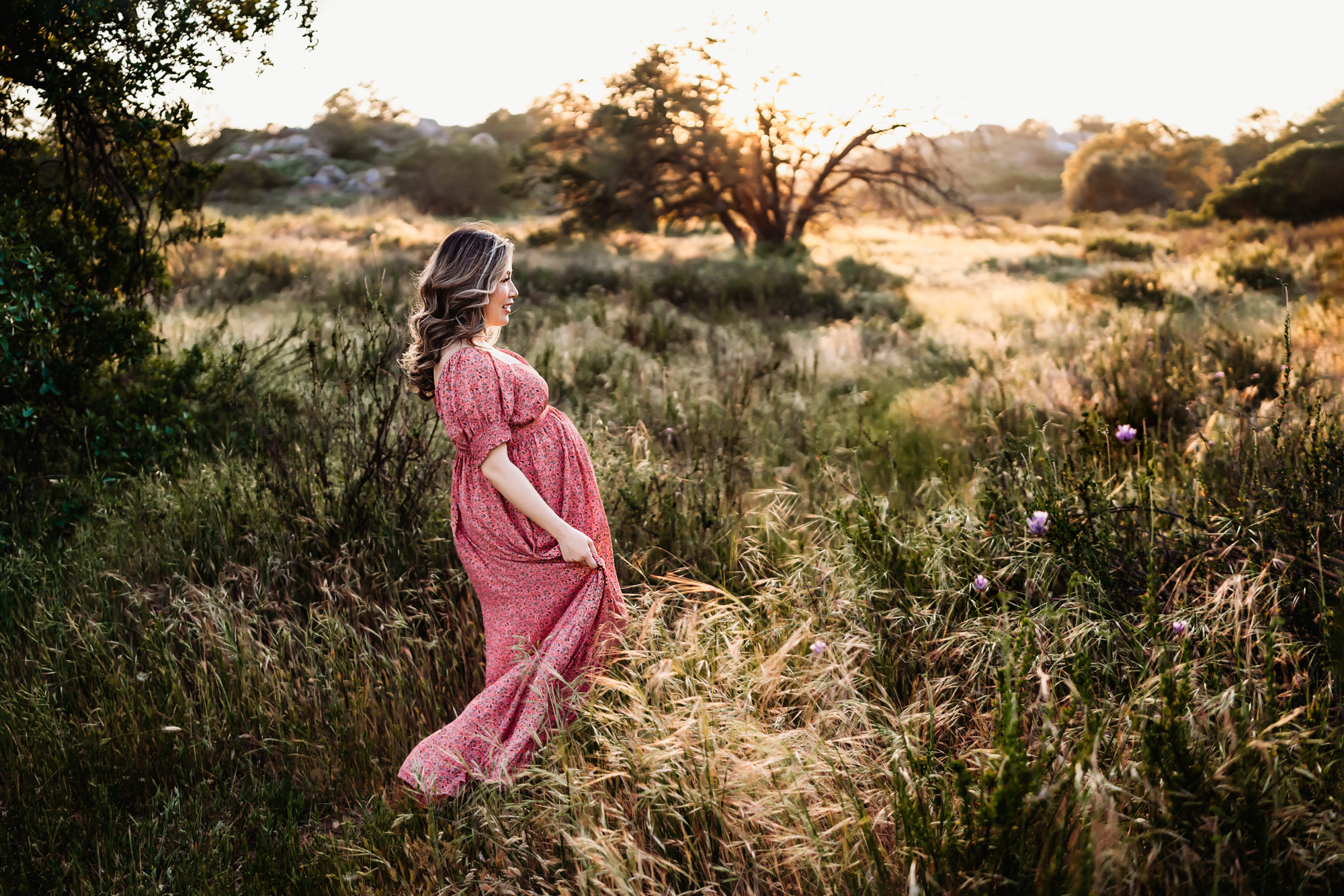 A pregnant woman wearing a pink dress walks through long grass in a field. This is during a San Diego lifestyle maternity photo session by Love Michelle Photography.